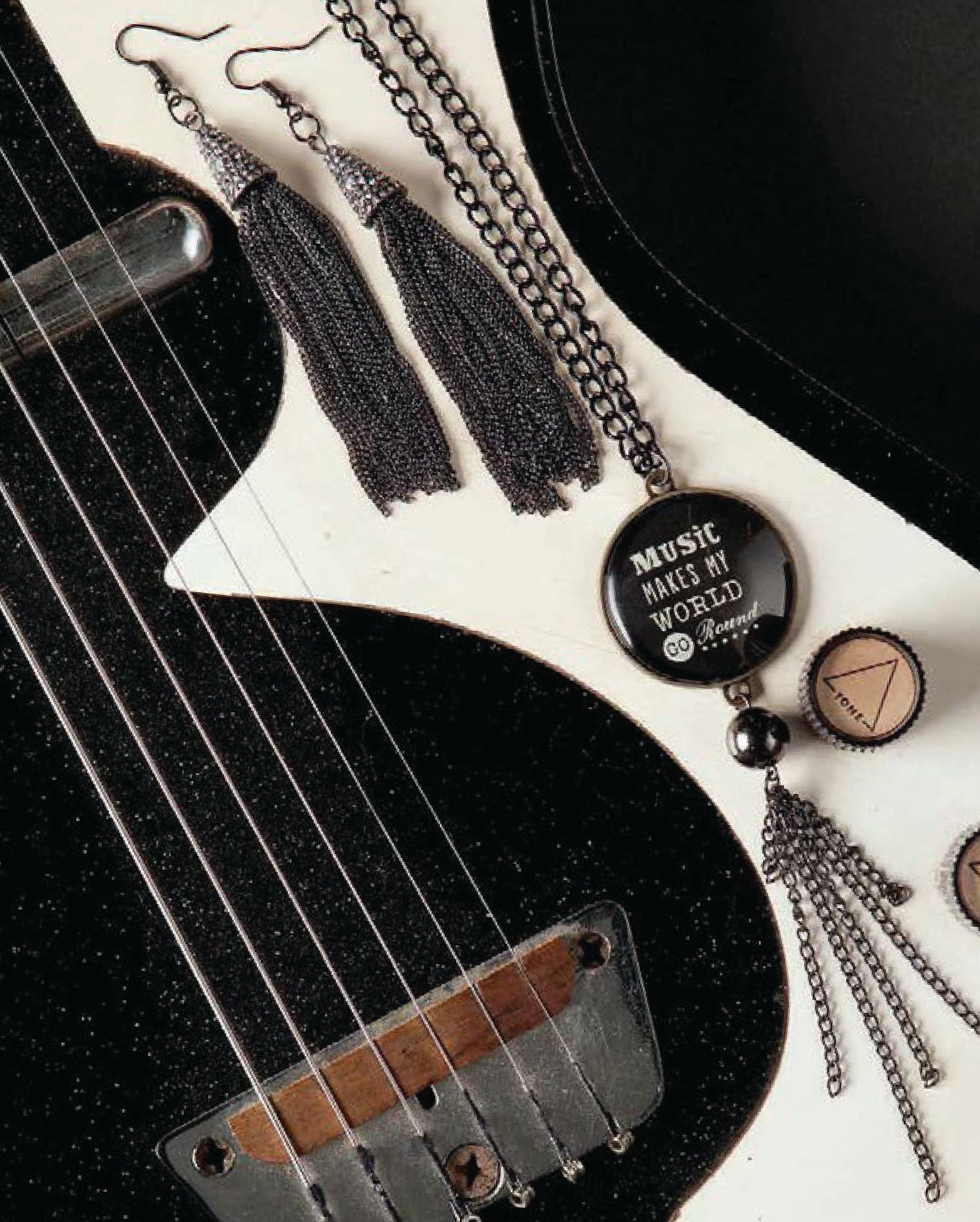 A close-up of black and white electric guitar with Wanderlust—Travel-Inspired Jewelry by Leisure Arts: black tassel earrings and a “Music makes my world go ‘round” necklace, perfect for music fans or travel jewelry lovers.