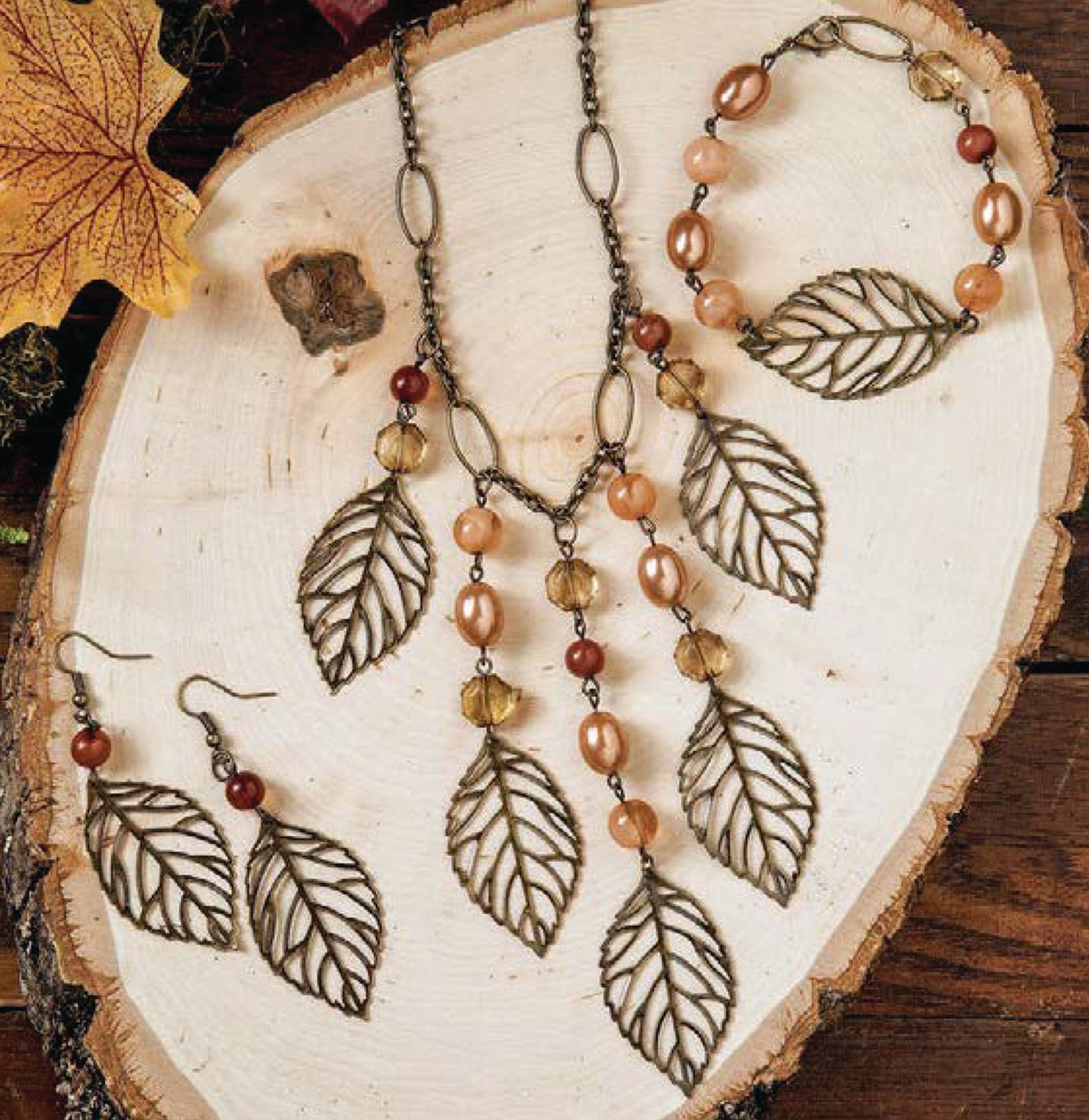 The Wanderlust—Travel-Inspired Jewelry set by Leisure Arts features a necklace, bracelet, and earrings with large metal leaf pendants and amber-brown beads, inspired by autumn leaves—a perfect mix-and-match accessory collection.
