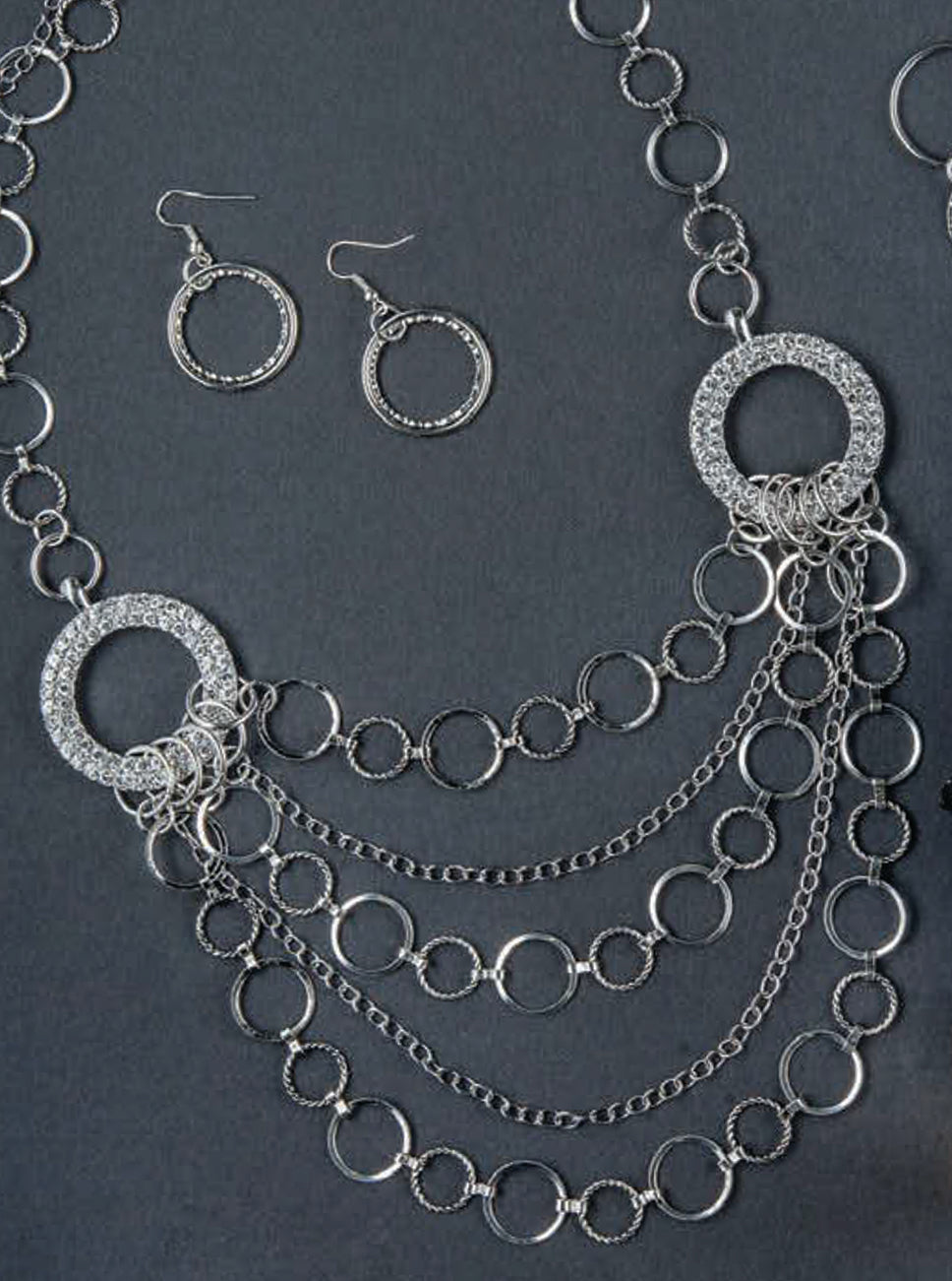 The Wanderlust—Travel-Inspired Jewelry set by Leisure Arts features a pair of silver hoop earrings and a stunning layered necklace composed of multiple interlocking circular links, expertly crafted with refined jewelry-making techniques and beautifully displayed against a dark background.