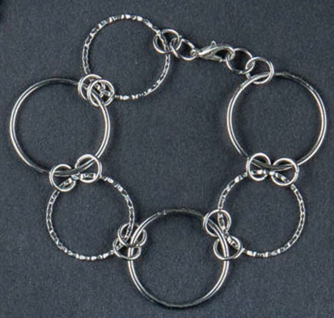 Introducing Wanderlust—Travel-Inspired Jewelry by Leisure Arts: a striking silver bracelet featuring six large interconnected rings, each joined by intricately twisted wire loops, and finished with a lobster clasp closure—an impressive showcase of jewelry-making techniques displayed against a dark background.