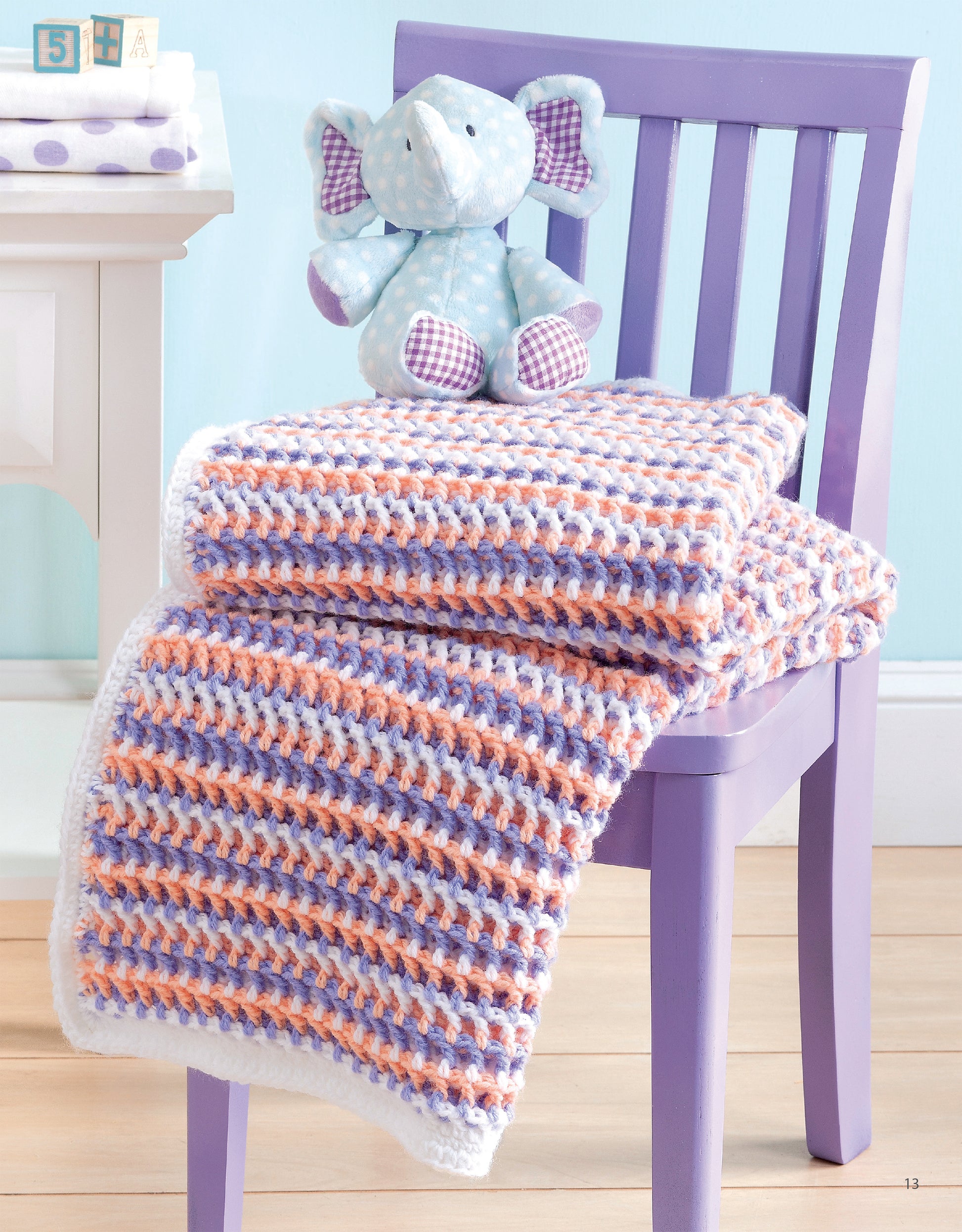 A pastel purple chair holds a folded Leisure Arts "Sweet Stripes for Baby" 3 Color Afghan with white, lavender, and peach stripes. A light blue stuffed elephant with checkered ears sits atop the blanket, displaying adorable baby afghan designs.
