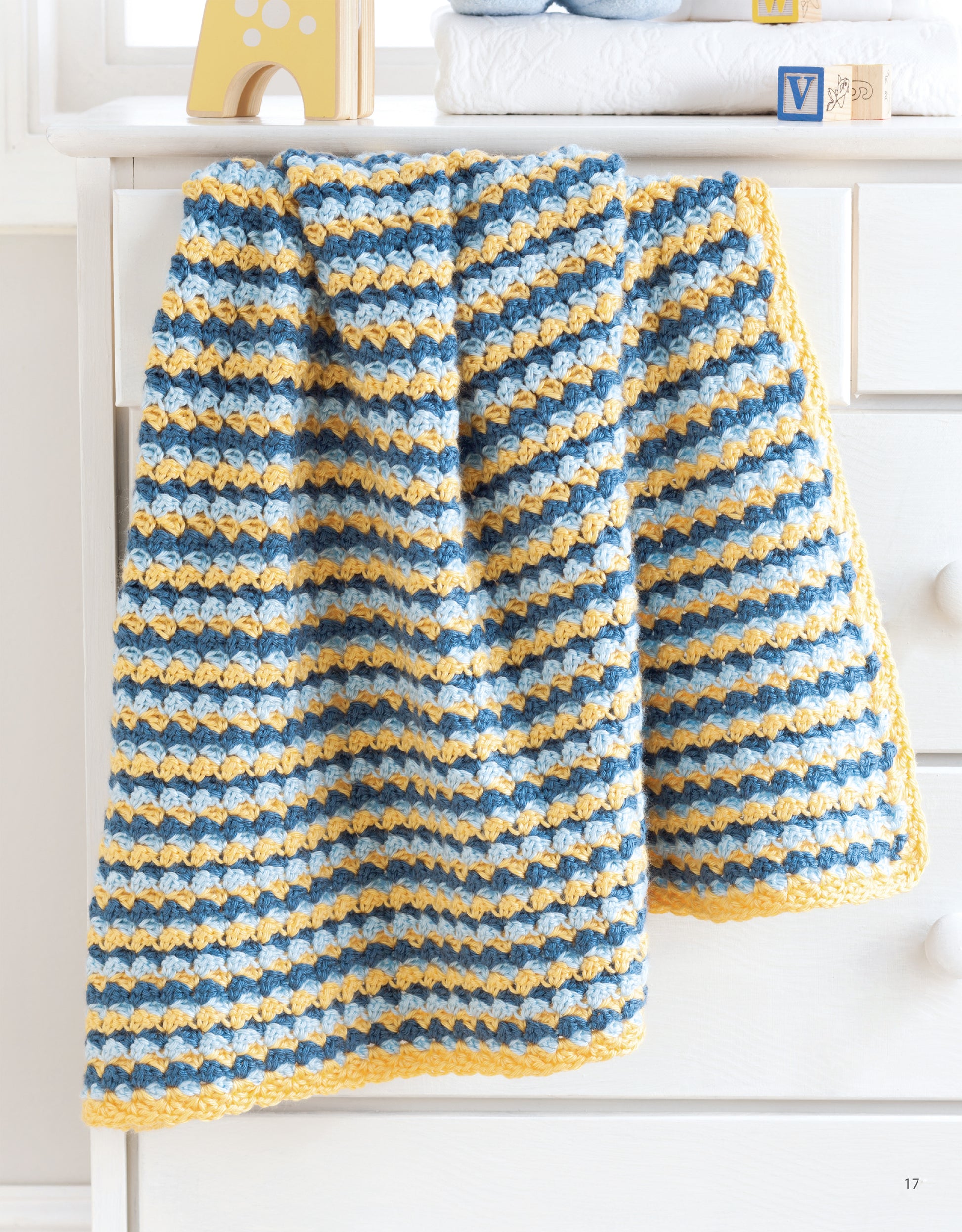 The “Sweet Stripes for Baby—Make 3 Color Afghans” blanket by Leisure Arts, featuring blue, yellow, and white stripes, is draped over a white dresser in a bright, softly lit room.