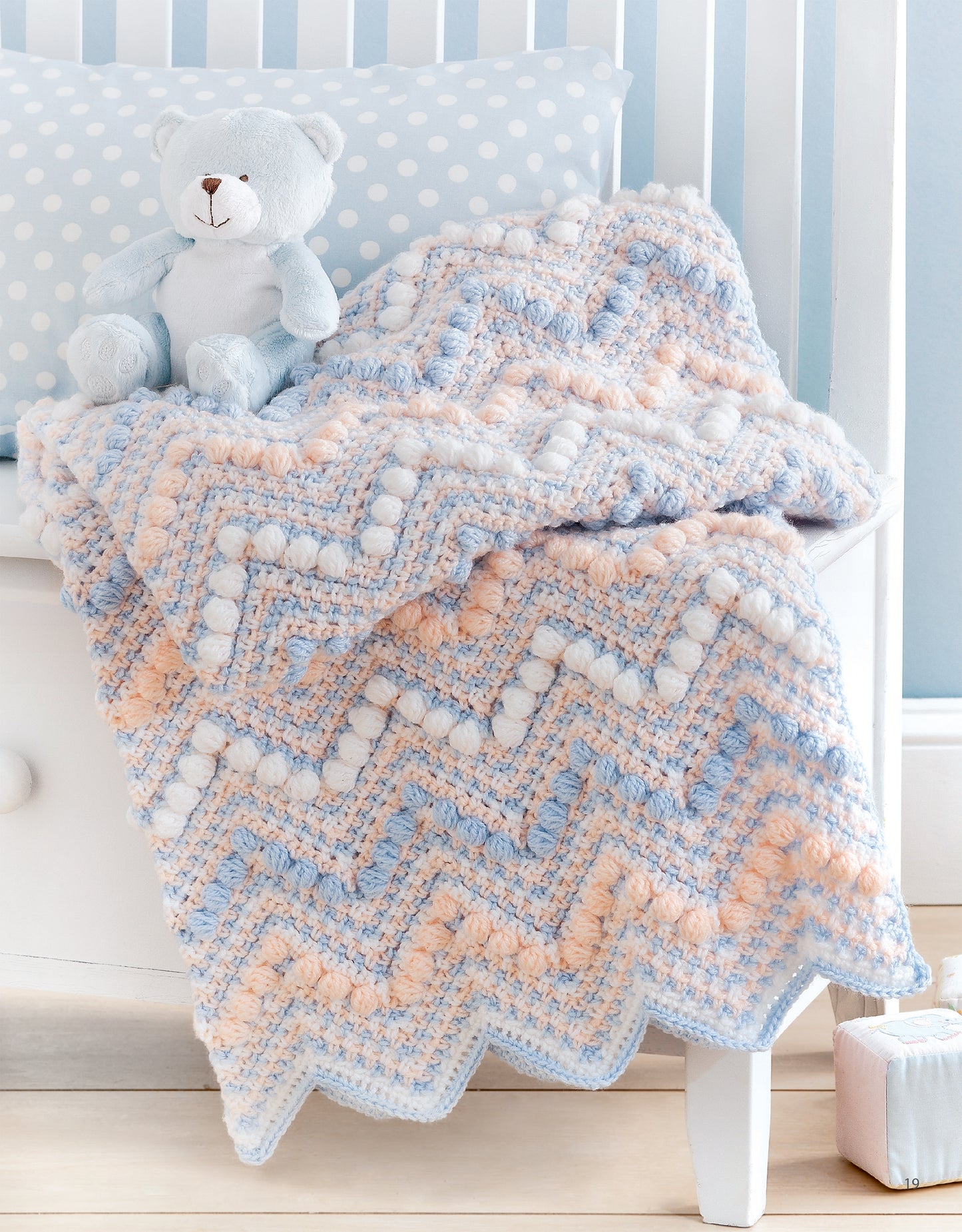 Sweet Stripes for Baby by Leisure Arts is a 3-color crocheted afghan in blue, peach, and white chevron with bobble stitches, draped over a white crib. Perfect for beginners seeking charming baby blanket inspiration. Blue teddy bear included for decor.