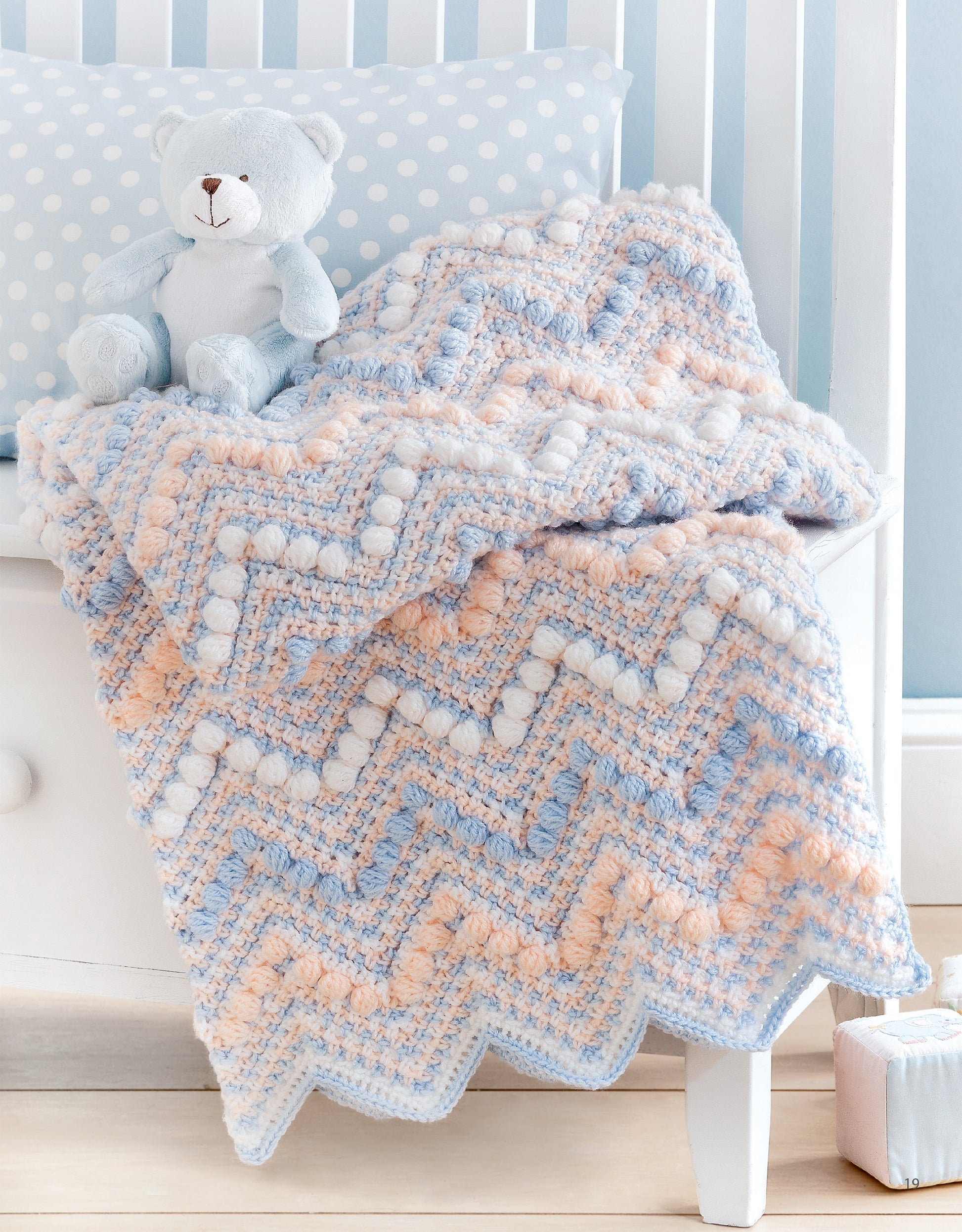 Sweet Stripes for Baby by Leisure Arts is a 3-color crocheted afghan in blue, peach, and white chevron with bobble stitches, draped over a white crib. Perfect for beginners seeking charming baby blanket inspiration. Blue teddy bear included for decor.