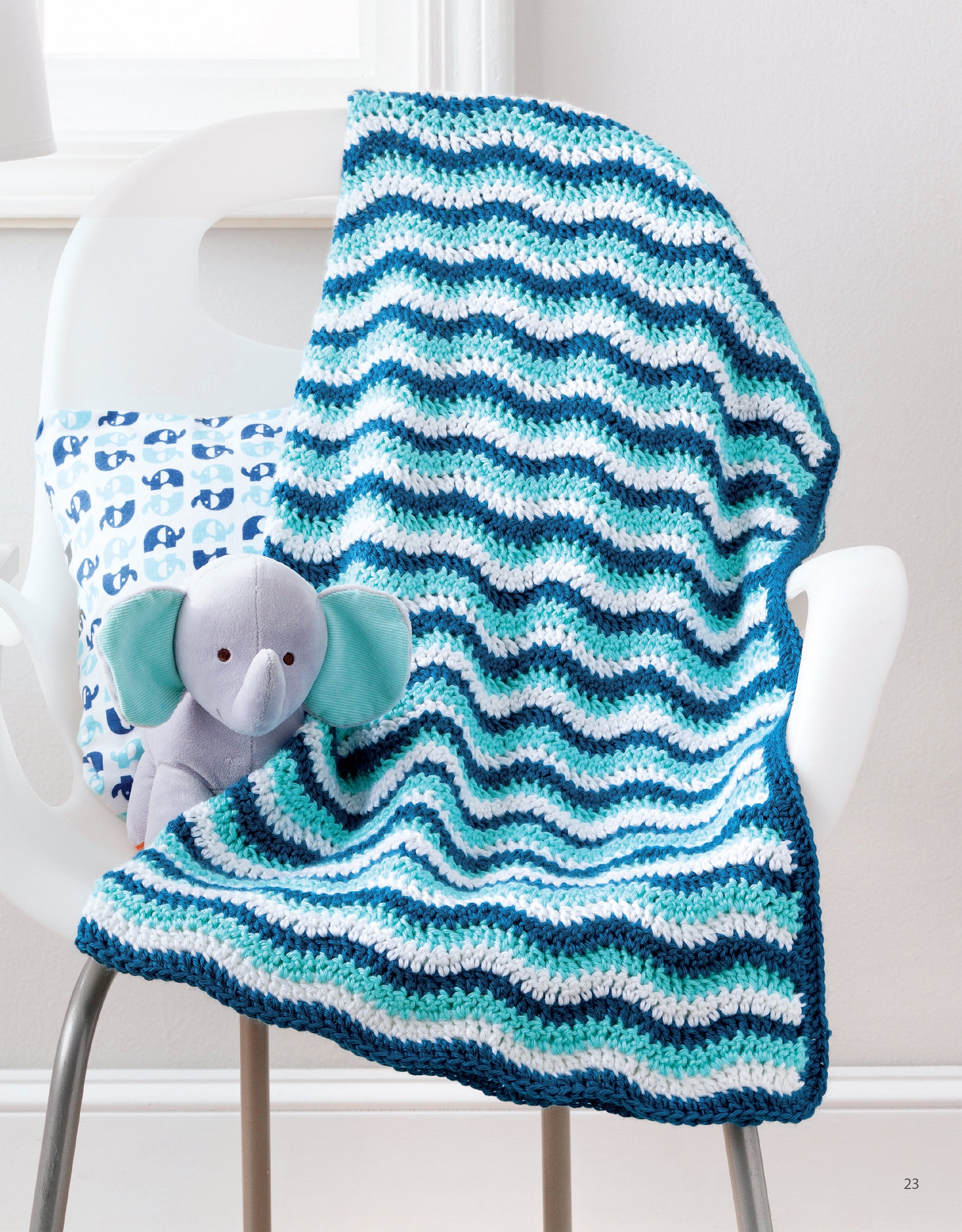 The Sweet Stripes for Baby—Make 3 Color Afghans blanket by Leisure Arts in blue, white, and teal stripes is draped over a white chair, next to a gray stuffed elephant and a pillow with blue whale prints.