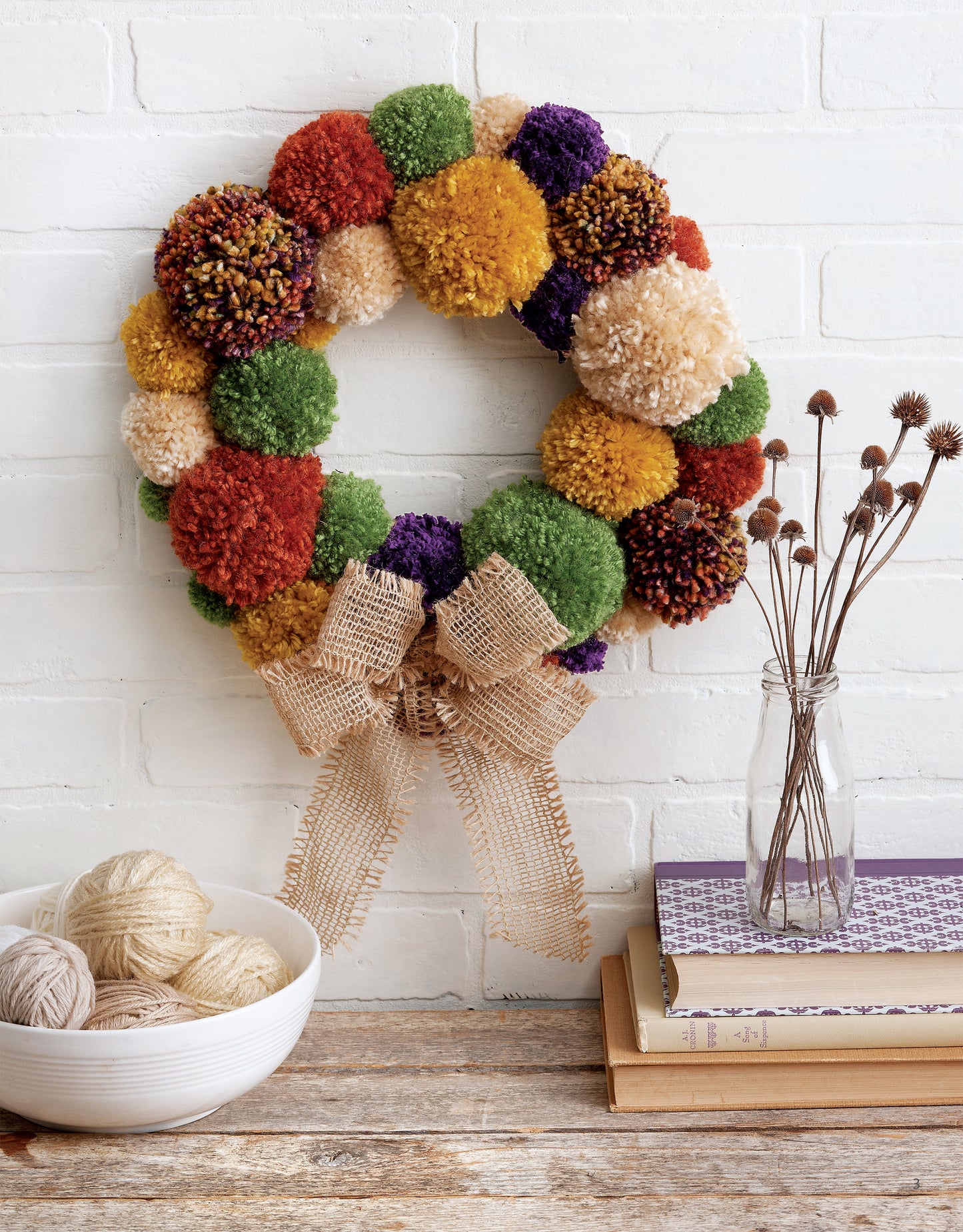 A vibrant pom-pom wreath with a burlap bow from Yarn Crafts by Leisure Arts hangs on a white brick wall, while a bowl of yarn balls and dried flowers in a glass vase accent stacked books on the wooden surface below.