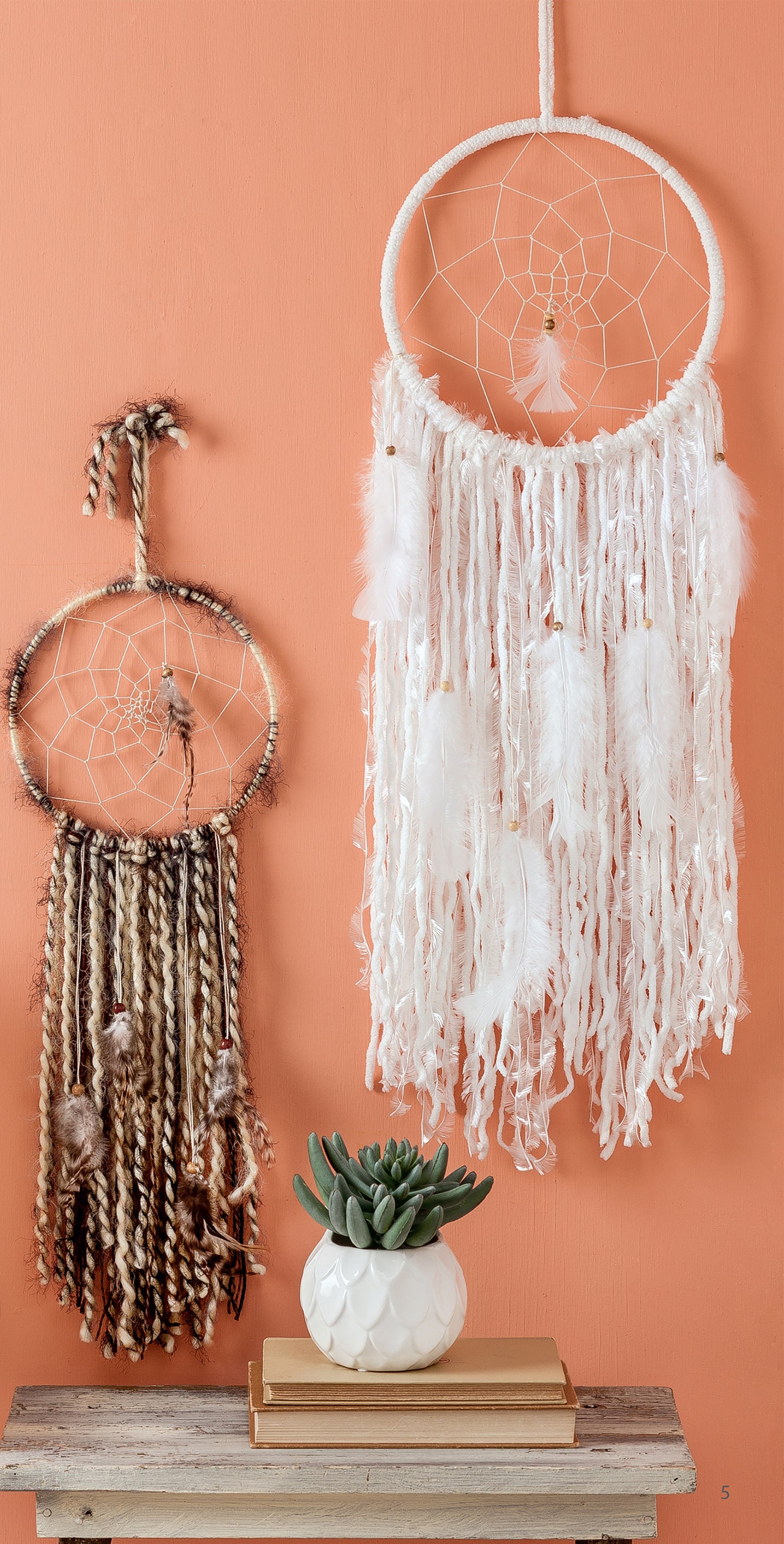 Two Yarn Crafts by Leisure Arts hang on a peach-colored wall above a small table with a white pot and green succulent, featuring feathers, woven details, and yarn-wrapped accents that add a cozy DIY touch.