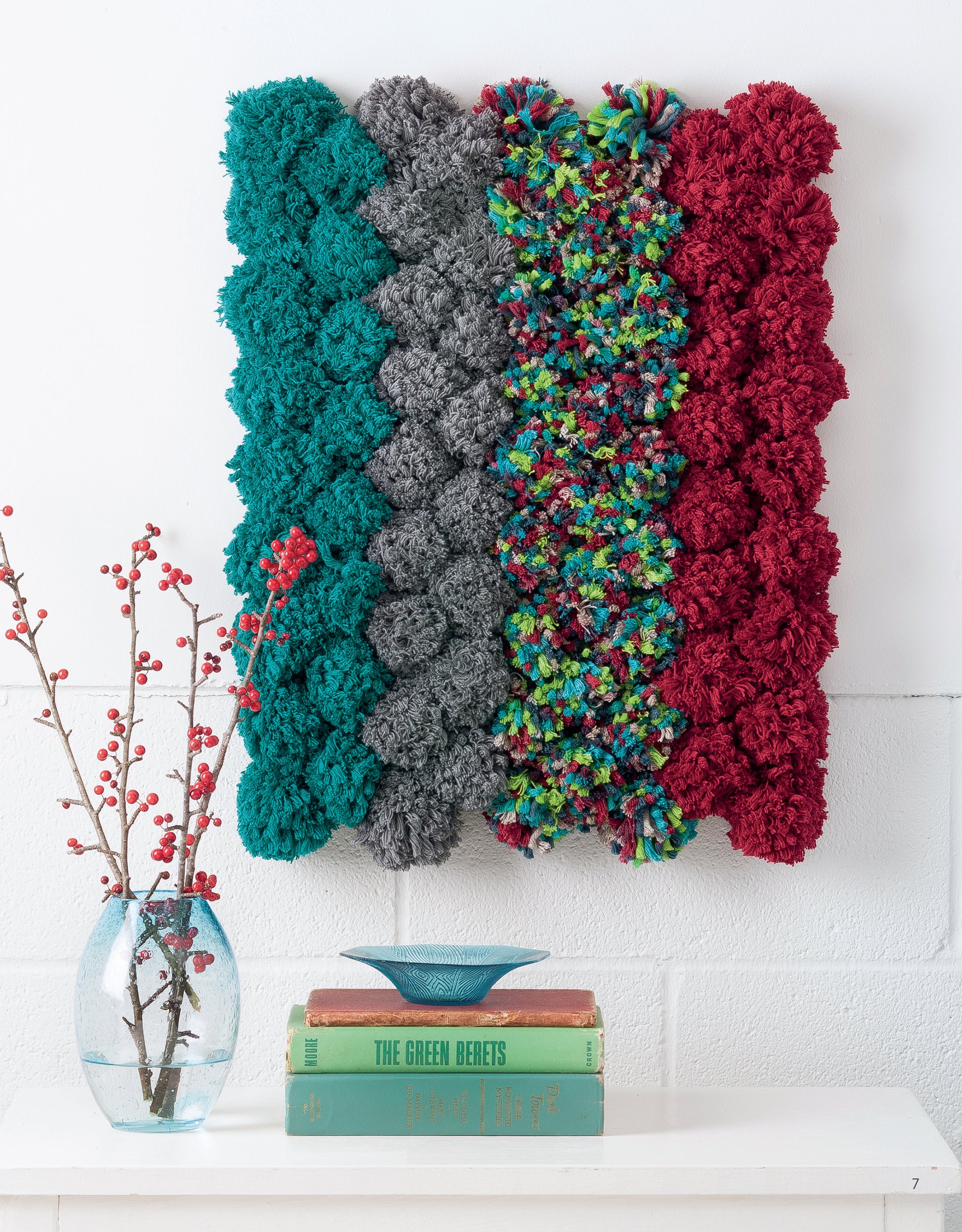 The Leisure Arts Yarn Crafts wall hanging features teal, gray, multicolored, and red textured yarn above a vase with red berries and books on a white shelf—perfect for creative DIY décor projects.
