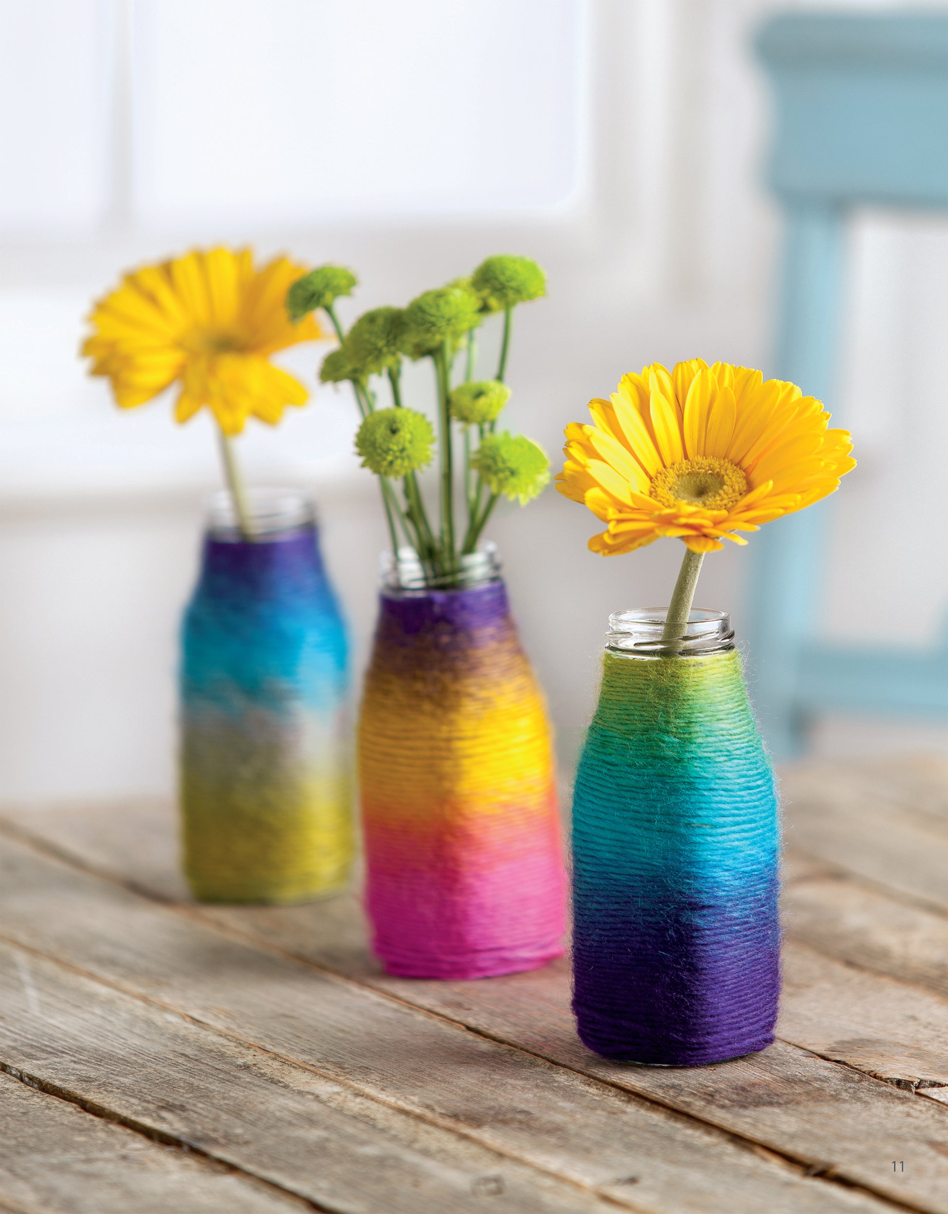 Leisure Arts' Yarn Crafts features three glass bottles decorated with yarn, holding yellow and green flowers. They rest on a rustic wooden table, backed by a blurred blue chair and white backdrop for a cozy DIY vibe.