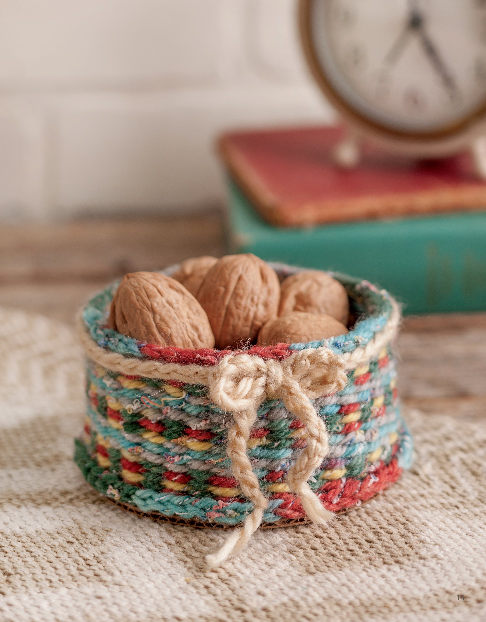 The Leisure Arts Yarn Crafts features a small, colorful woven basket with a yarn bow holding whole walnuts, perfect for DIY projects. A blurred clock and book in the background create a cozy, rustic vibe.
