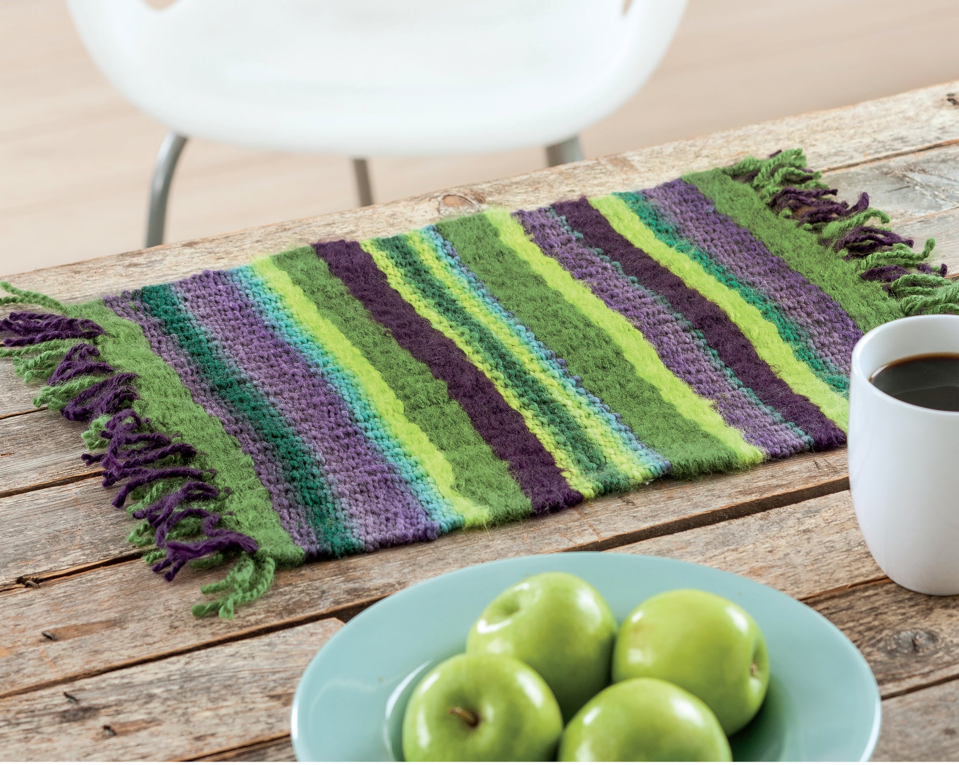 The Leisure Arts Yarn Crafts striped handwoven placemat in green, blue, and purple sits on a rustic wooden table with a white mug, blue plate holding green apples, and a white chair in the background.
