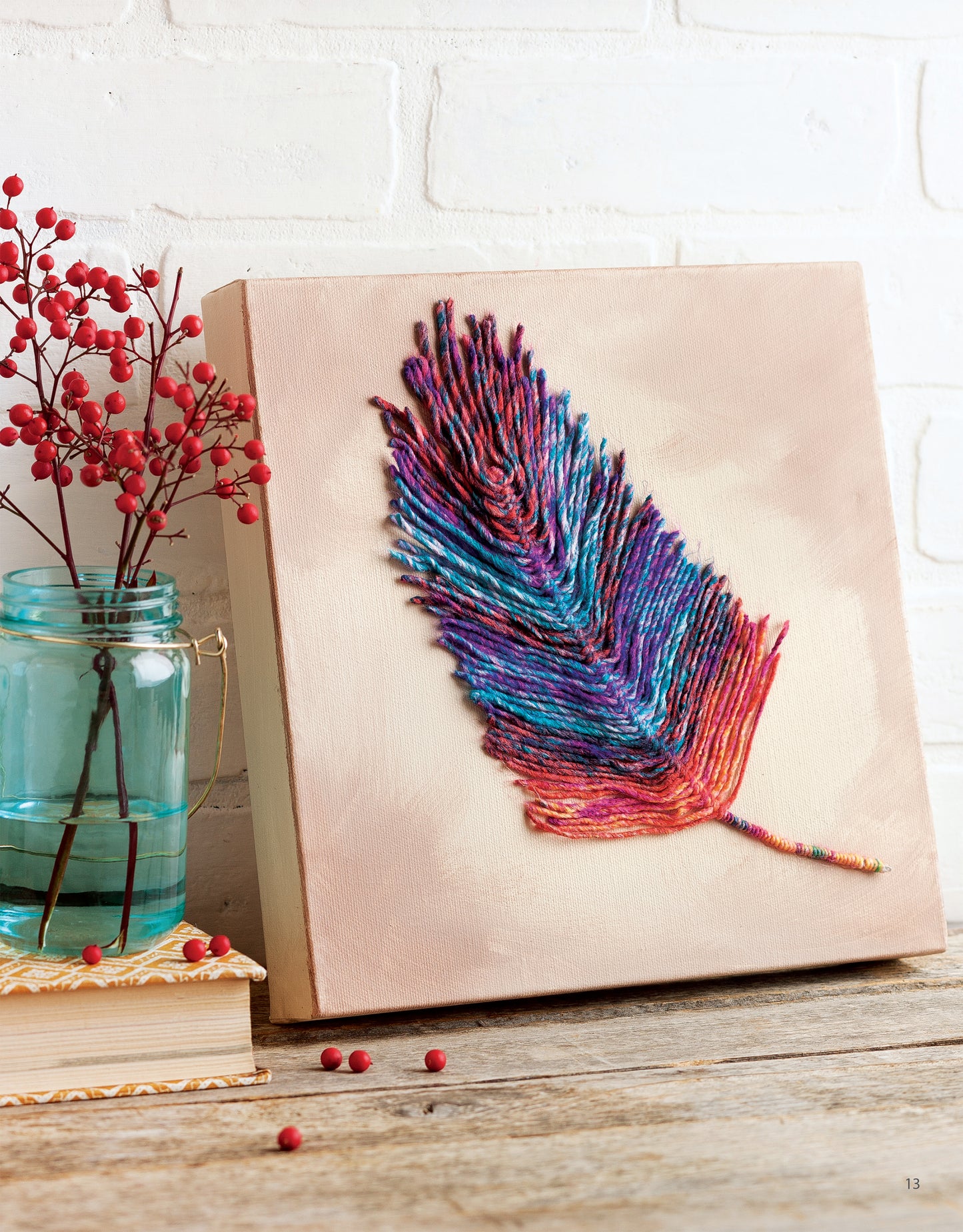 The Yarn Crafts art piece by Leisure Arts displays a colorful feather on canvas, set on a wooden surface near a blue mason jar with red berries and books, all against a white brick wall.