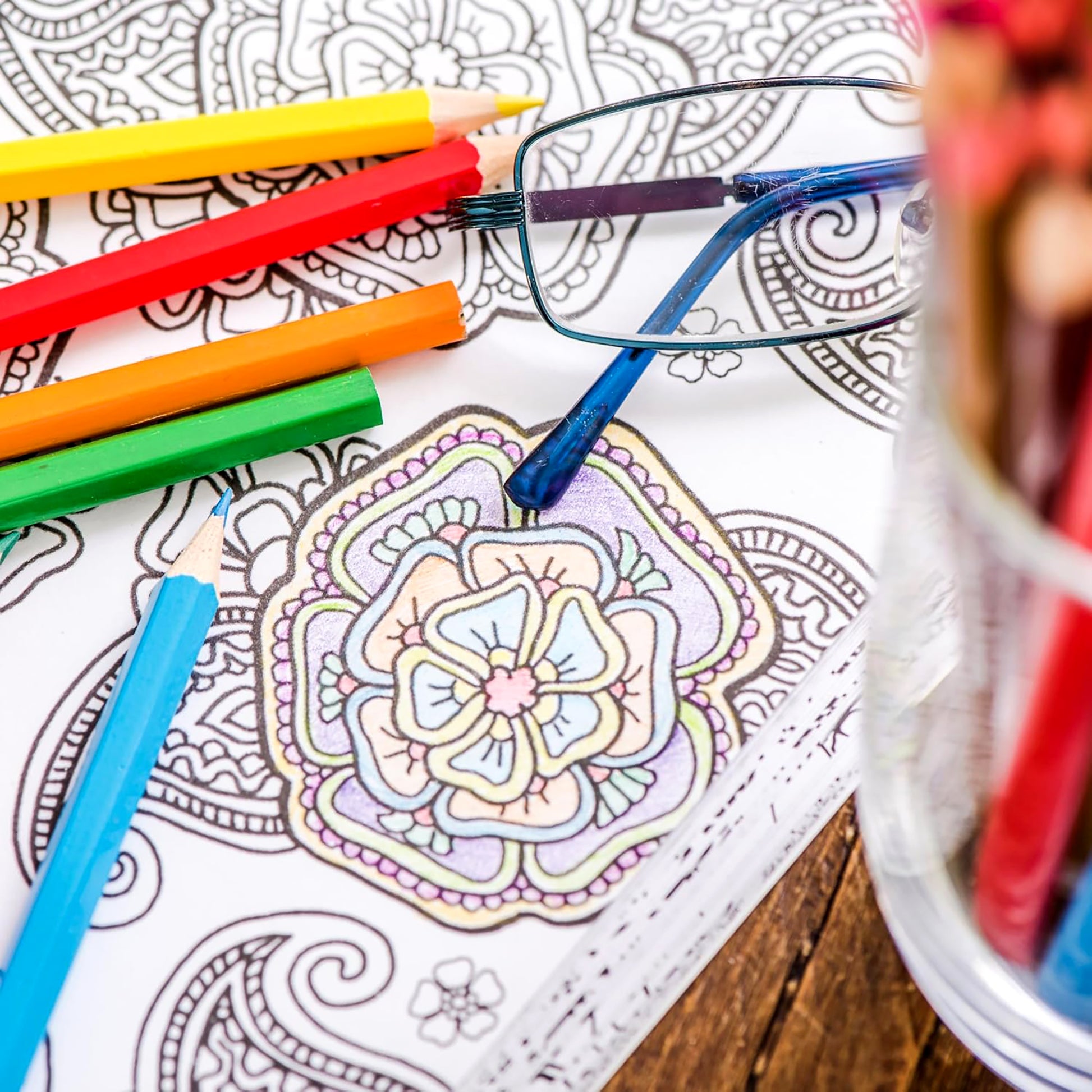 A close-up of a partially colored floral design in "The Best of Color Art for Everyone" by Leisure Arts, with colored pencils, glasses, and a pencil container on a wooden surface—perfect for stress relief.