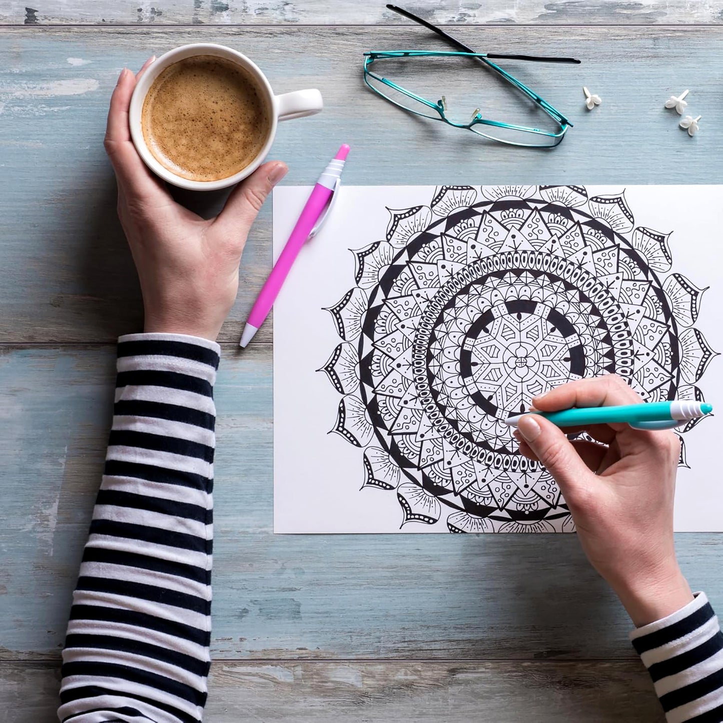 Wearing a striped shirt, someone colors an intricate mandala from "The Best of Color Art for Everyone" by Leisure Arts, coffee cup in hand; glasses, earbuds, and markers on a blue table create a relaxing, stress-relieving scene.