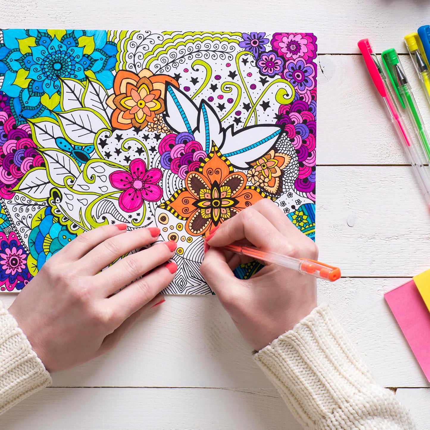 Wearing a cream sweater, someone colors an intricate floral page from Leisure Arts’ "The Best of Color Art for Everyone" with an orange pen. Colored pens and sticky notes rest on the white wooden table, setting a calm, stress-relieving mood.