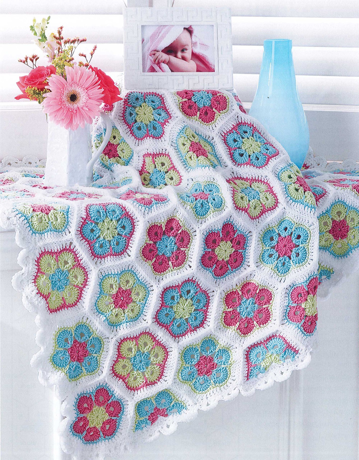 The Crochet Sweet Baby Blankets by Leisure Arts, featuring vibrant hexagonal floral motifs, is draped over a white table with pink flowers in a vase, a blue vase, and a framed baby photo nearby.