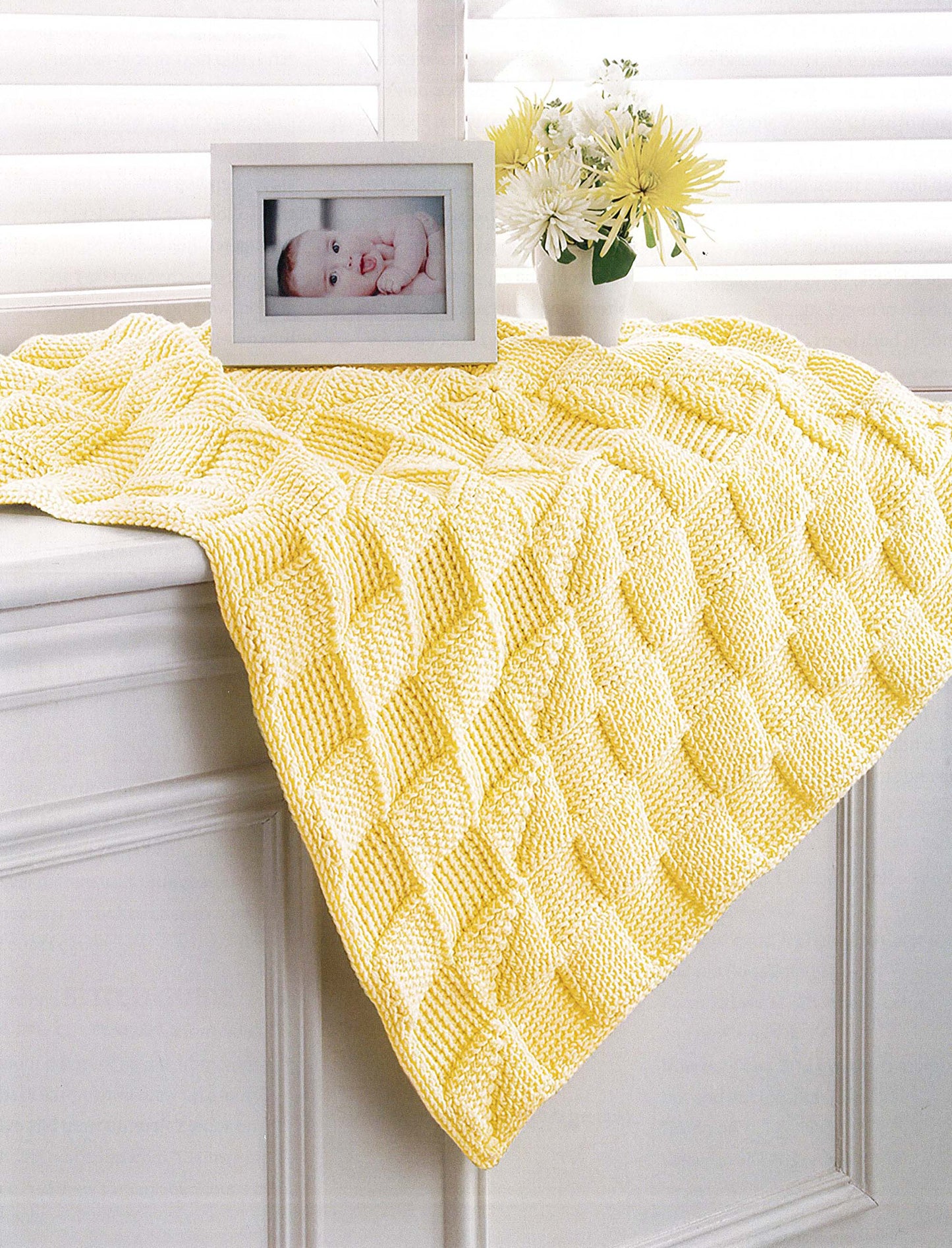 The Crochet Sweet Baby Blankets by Leisure Arts in soft yellow and white are draped over a windowsill next to yellow and white flowers and a framed baby photo, with white blinds in the background.