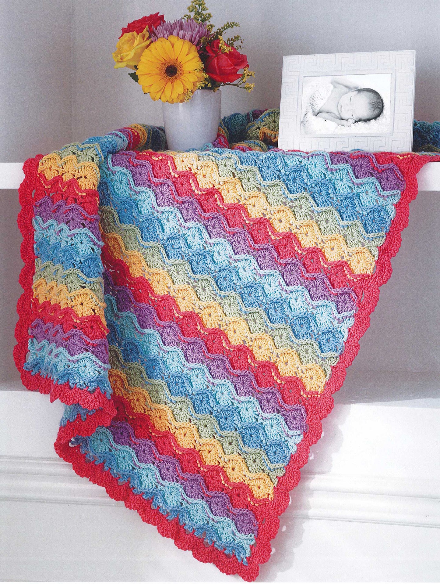 The Crochet Sweet Baby Blankets by Leisure Arts, featuring wavy blue, yellow, pink, and red stripes, is draped over a white shelf decorated with a flower vase and a framed black-and-white baby photo.