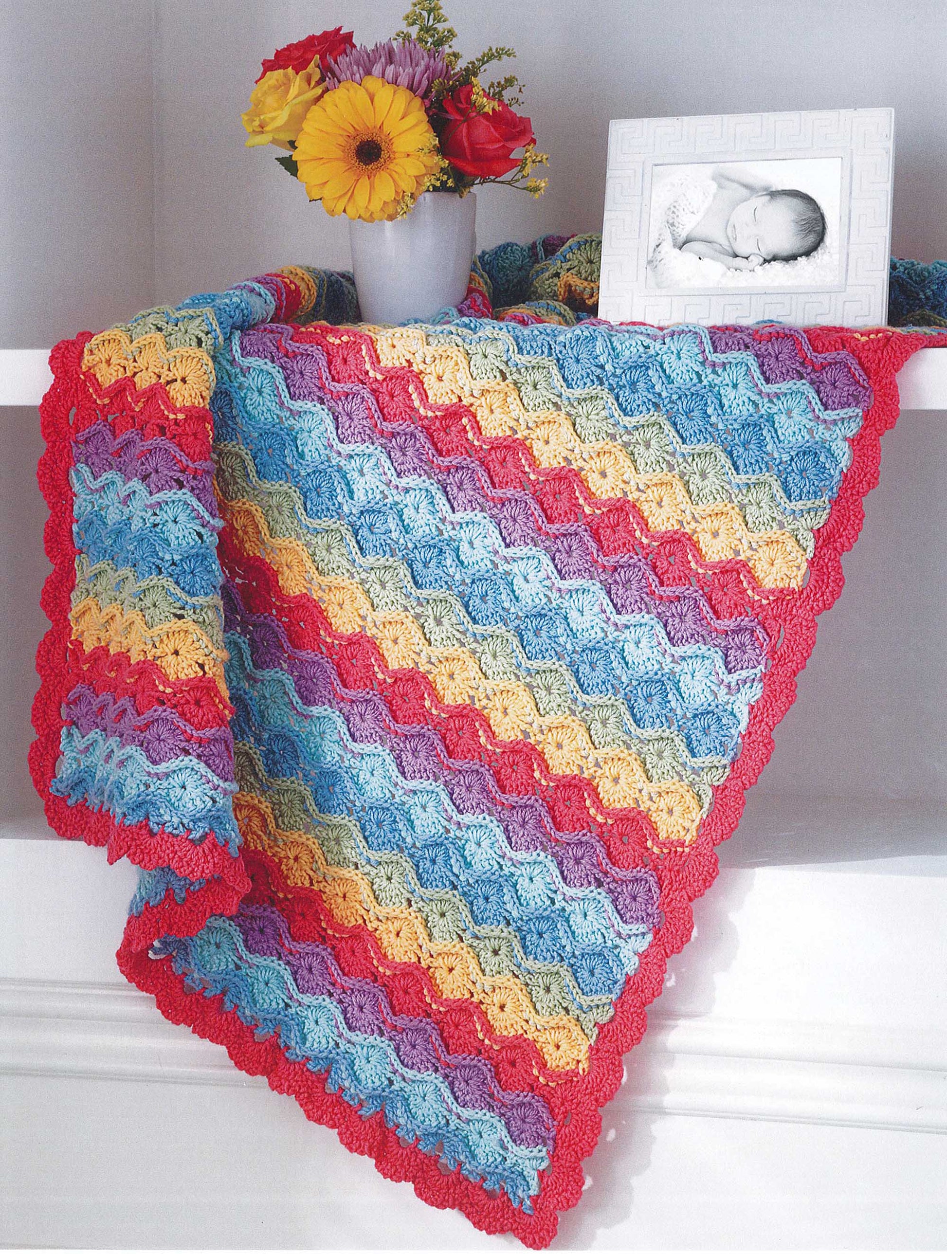 The Crochet Sweet Baby Blankets by Leisure Arts, featuring wavy blue, yellow, pink, and red stripes, is draped over a white shelf decorated with a flower vase and a framed black-and-white baby photo.