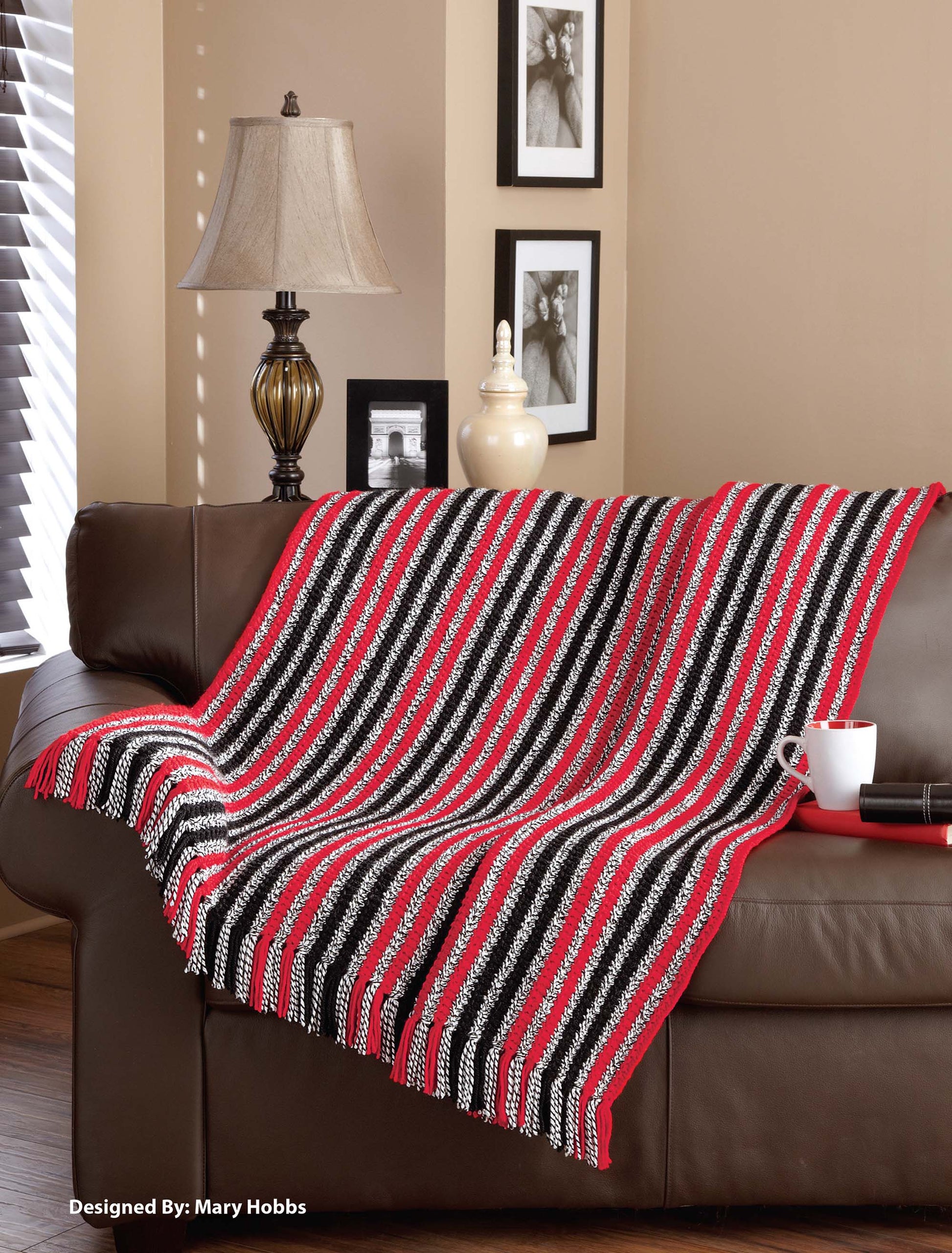 A cozy Knit Classic Afghans by Leisure Arts, featuring red, black, and white stripes, is draped over a brown sofa. A mug and book rest on the arm, with framed photos and a lamp in the background adding inviting charm.