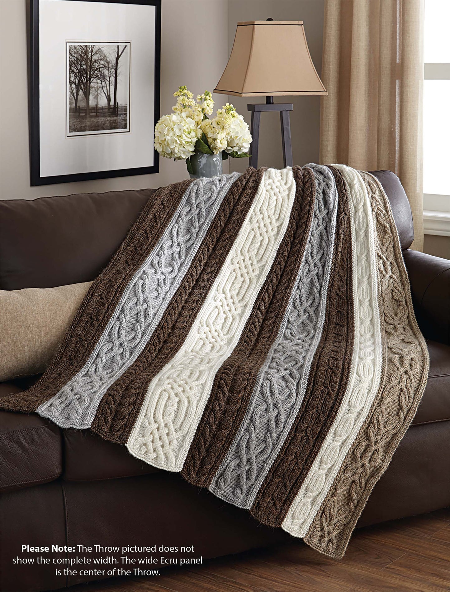 The Knit Classic Afghans by Leisure Arts, featuring textured cable patterns in brown, gray, and cream stripes, adds cozy charm to your home draped over a brown couch. A lamp, white flowers, and framed art decorate the table behind it.