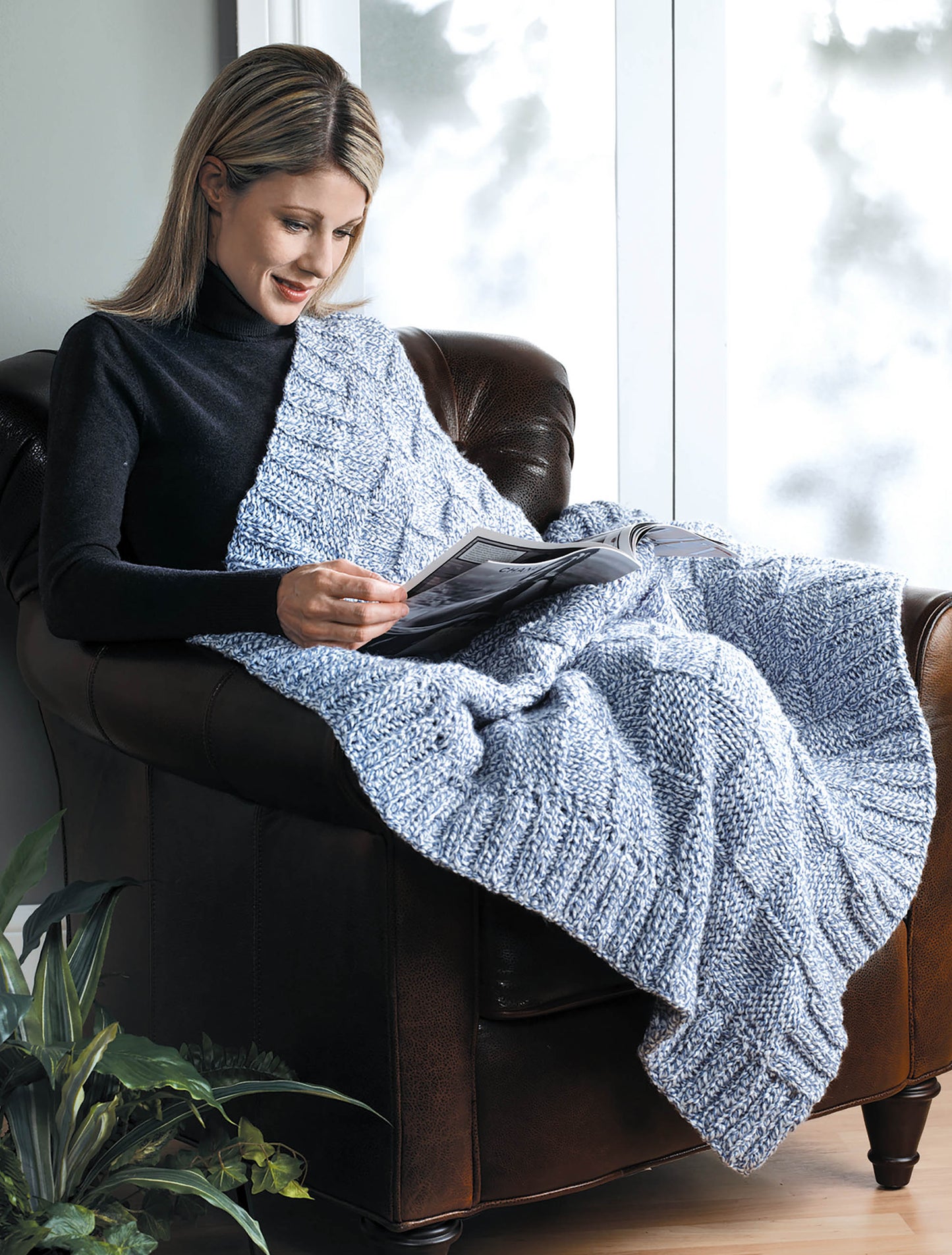 A woman with blonde hair relaxes in a brown armchair by a window, wrapped in a light blue Knit Classic Afghans throw from Leisure Arts. A green potted plant beside her enhances the cozy decor as she reads a magazine.