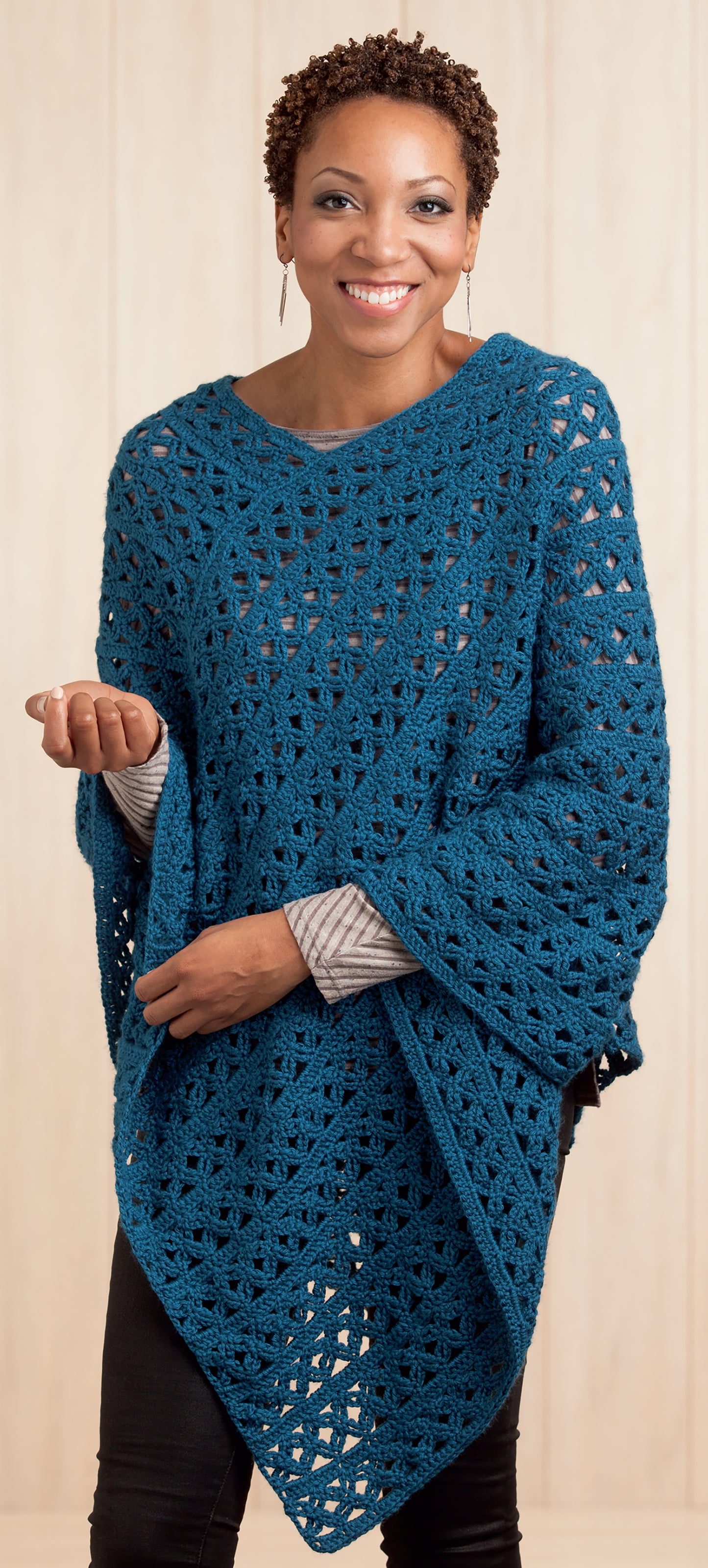 A smiling person models the Leisure Arts Boho Chic Crochet Poncho, a blue beginner-level design, over a gray shirt and black pants against a light background, standing with one hand raised slightly.