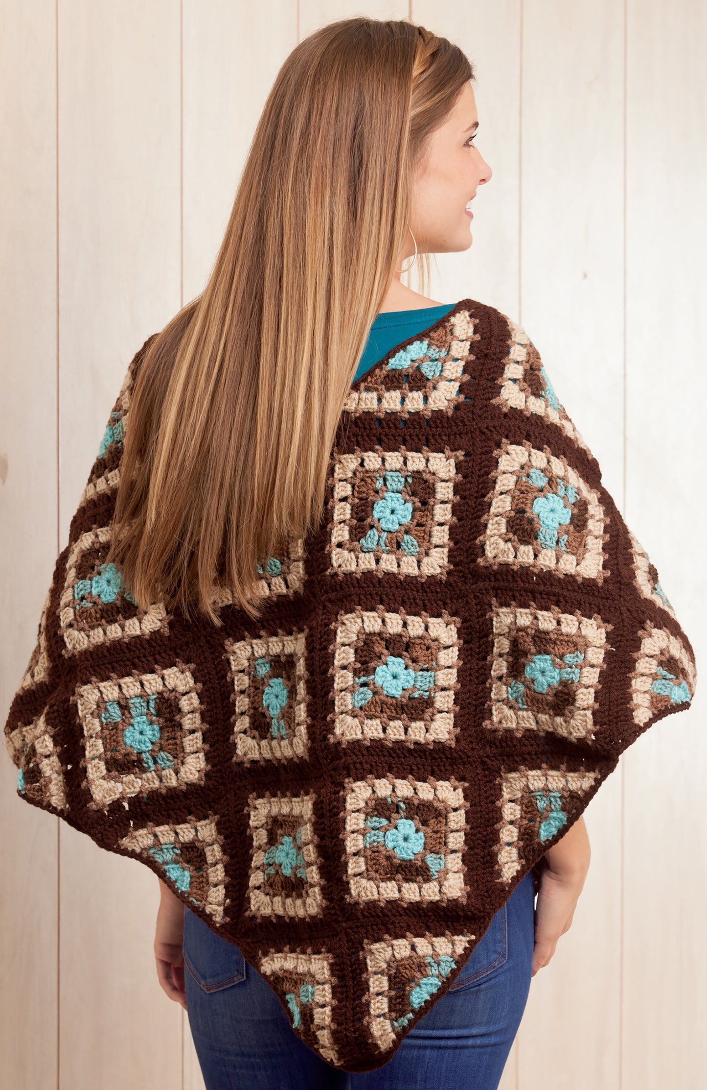 A woman with long straight hair models the Boho Chic Crochet Ponchos by Leisure Arts, featuring brown, tan, and teal granny squares—perfect for fans of easy crochet designs—standing with her back to a wooden background.