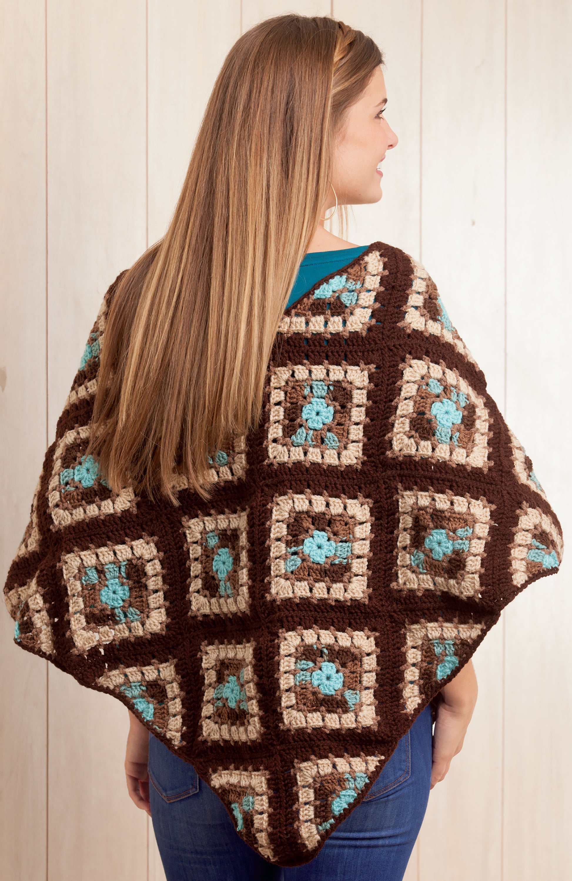 A woman with long straight hair models the Boho Chic Crochet Ponchos by Leisure Arts, featuring brown, tan, and teal granny squares—perfect for fans of easy crochet designs—standing with her back to a wooden background.