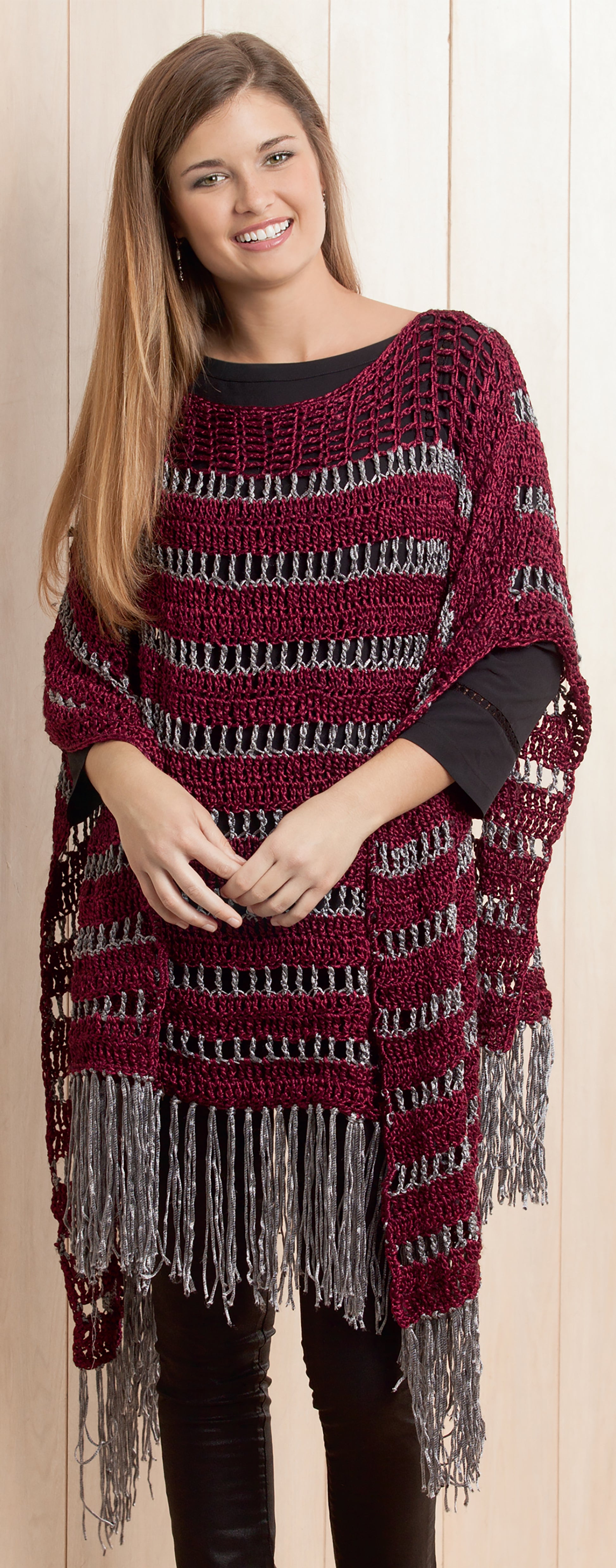 A woman with long brown hair smiles at the camera, wearing the Boho Chic Crochet Poncho by Leisure Arts in burgundy and gray stripes with fringe, standing before light wooden panels.