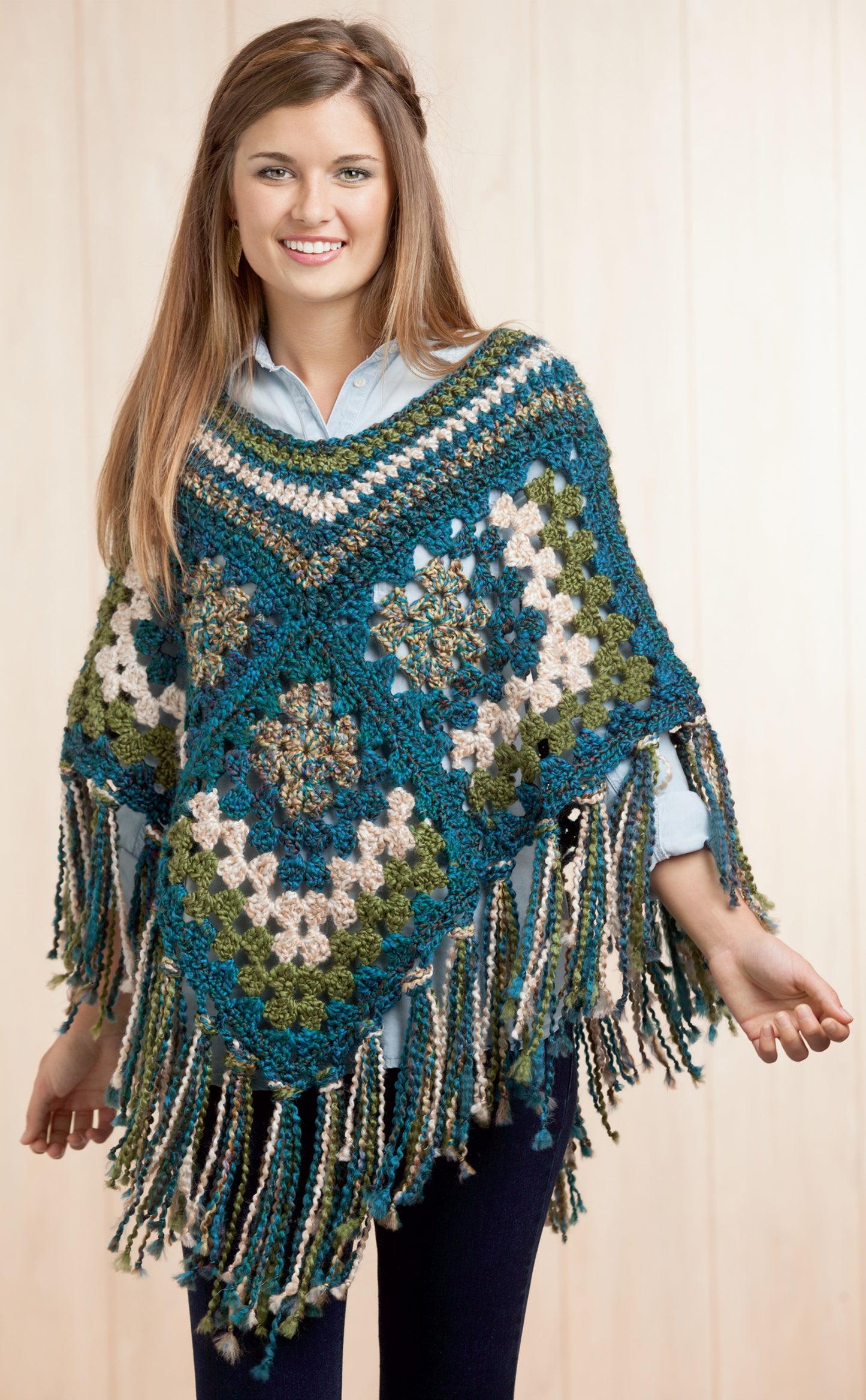 A smiling woman with long brown hair wears the Boho Chic Crochet Ponchos by Leisure Arts, featuring colorful blue, green, beige, and white granny squares with long fringe. She stands against a light wood background.
