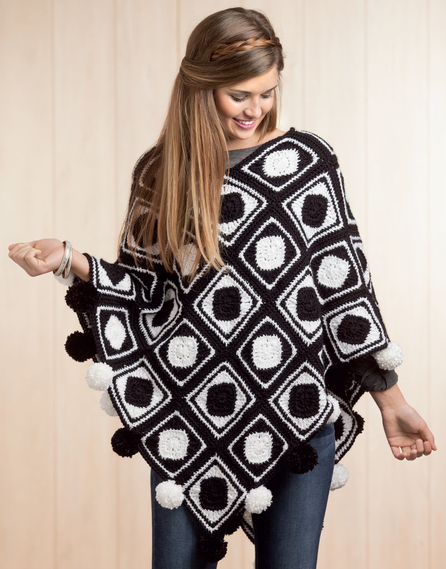 A smiling woman with long hair models the Boho Chic Crochet Ponchos by Leisure Arts, featuring a black and white geometric pattern with pom-poms, paired with blue jeans. She stands against a light wood background, looking down to her left.