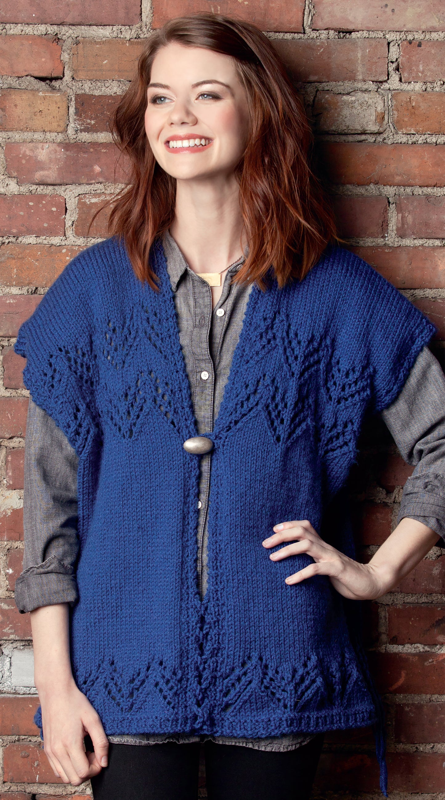 A smiling woman with shoulder-length brown hair stands before a brick wall, wearing a gray shirt and the Knit Poncho Rebound by Leisure Arts—a royal blue knitted wrap fastened with a round button. She poses confidently with one hand on her hip.