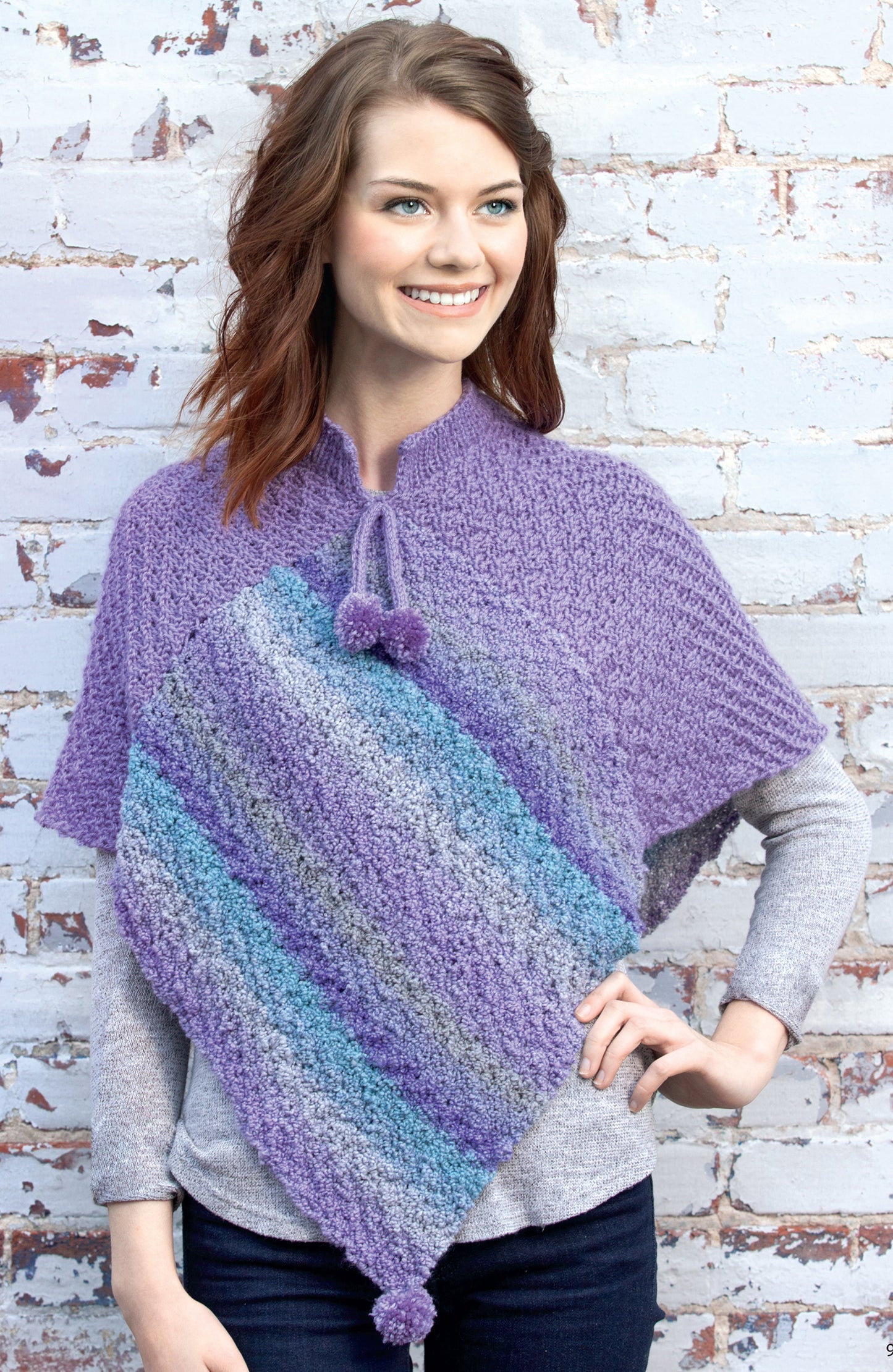 A woman smiles in front of a white brick wall, wearing the Knit Poncho Rebound by Leisure Arts—a textured purple poncho with blue and gray diagonal stripes, accented with pom-poms at the neckline and hem.