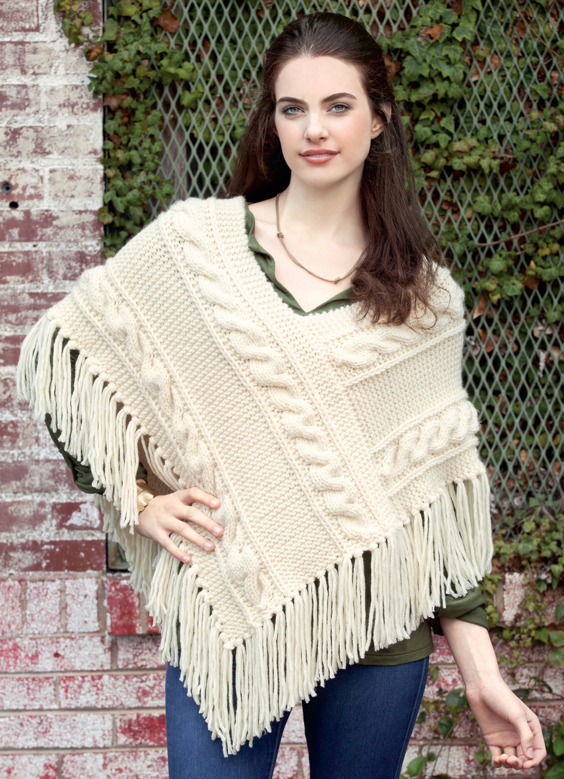 A woman with long brown hair models the Knit Poncho Rebound by Leisure Arts—a cream cable knit wrap with fringe—over a green blouse and blue jeans as she stands by a brick wall with green vines, hand on hip.