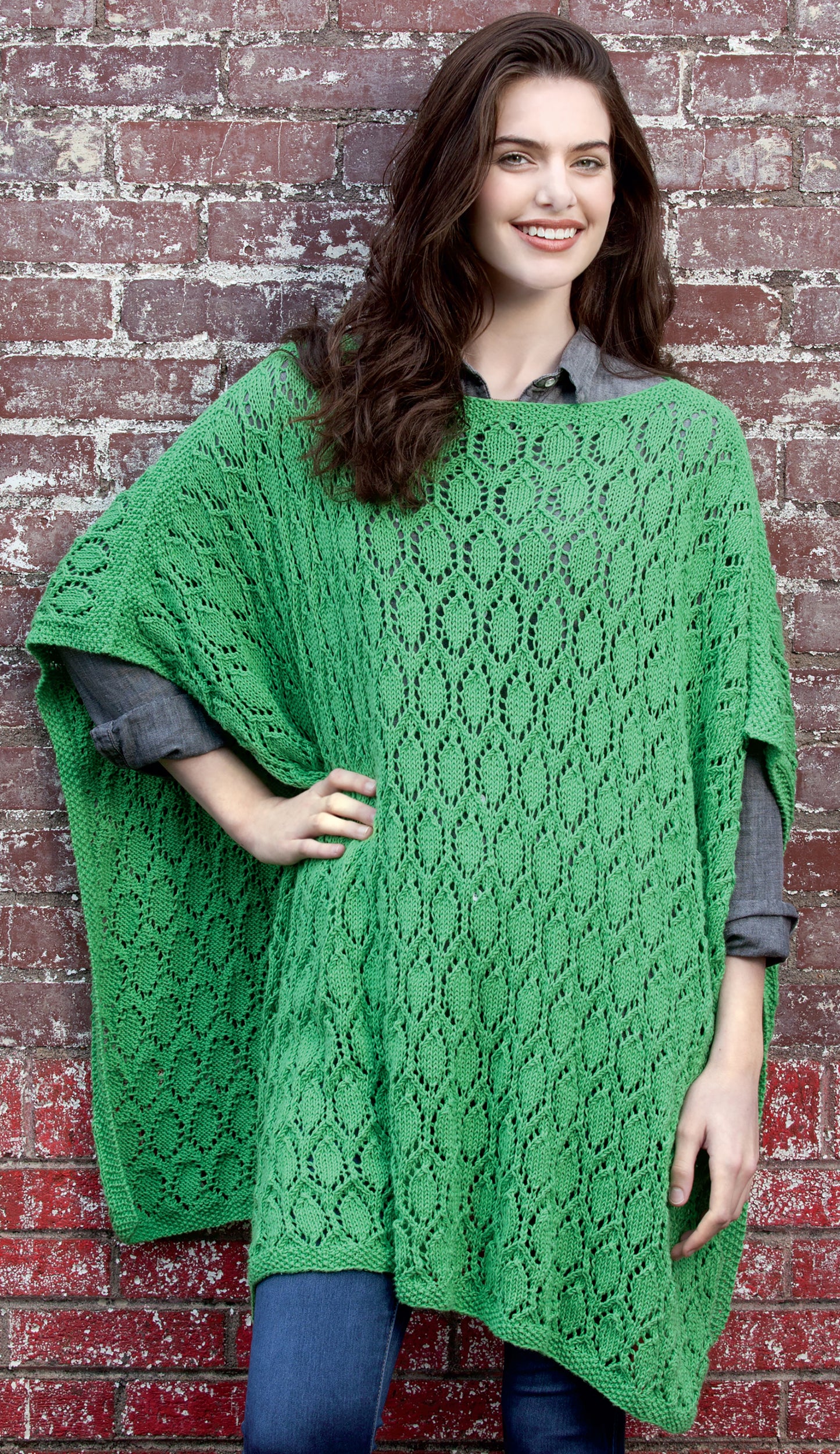 A woman stands in front of a brick wall, smiling and wearing the Knit Poncho Rebound by Leisure Arts over a gray shirt and blue jeans. Her right hand rests on her hip, and her dark hair falls loosely over her shoulders.