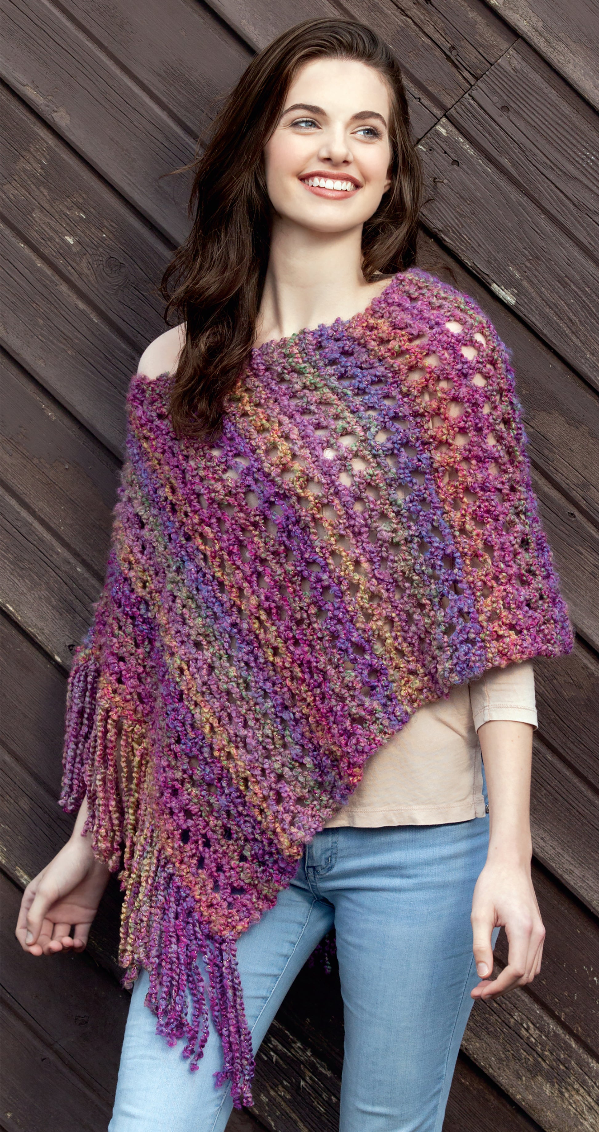 A woman leans against a wooden wall, smiling in the Leisure Arts Knit Poncho Rebound—a multicolored, textured wrap in purple, pink, and orange with fringe—styled over a beige top and light blue jeans.