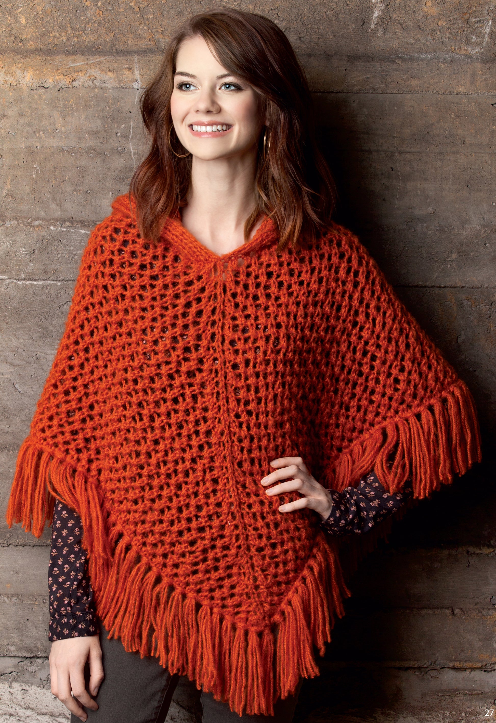 A woman with brown hair smiles while wearing the Leisure Arts Knit Poncho Rebound, a chunky orange crocheted wrap with fringe, standing against a textured brown wall.