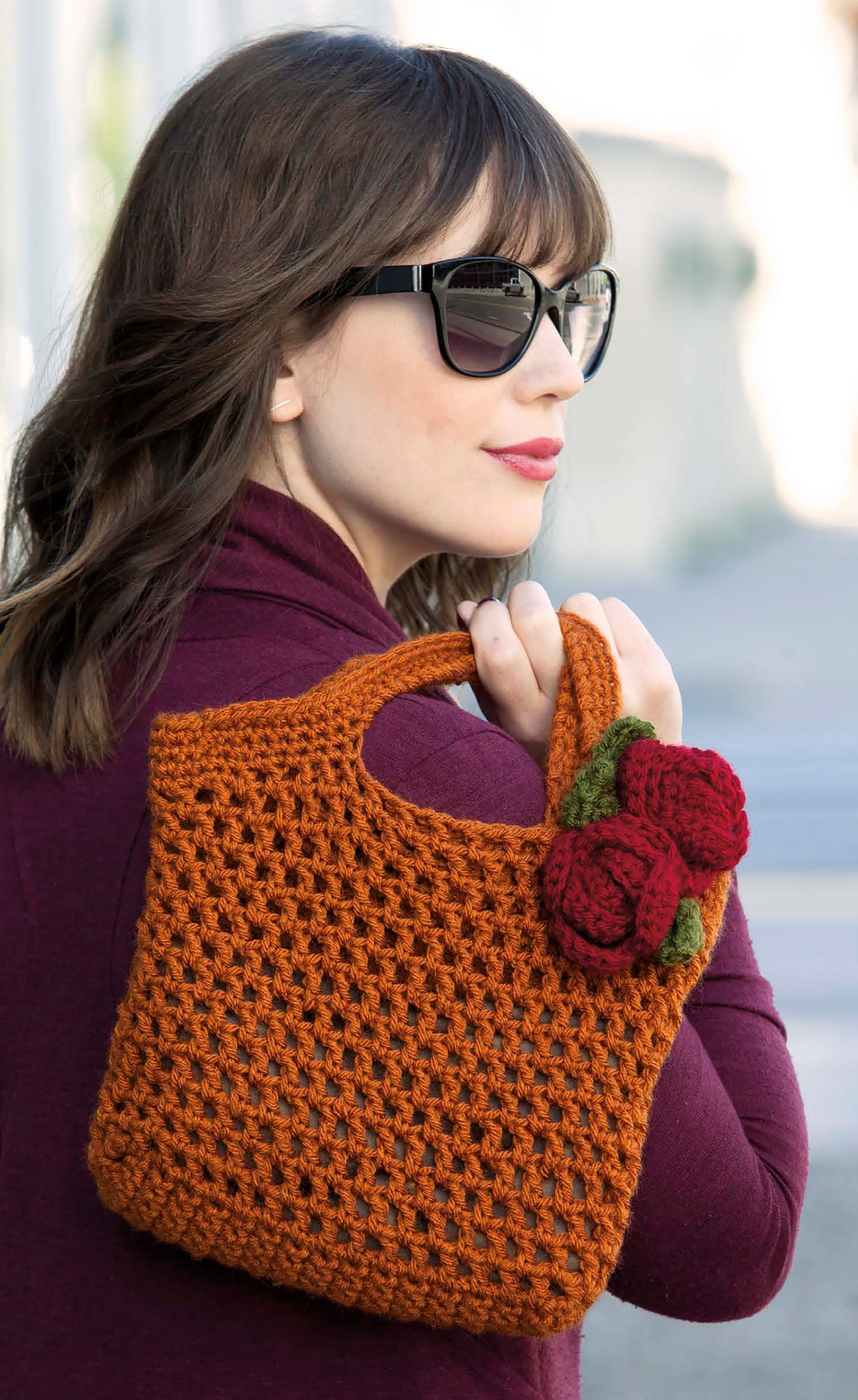 A woman in sunglasses and a burgundy top carries the Easy Essential Bags Crochet by Leisure Arts—a brown crocheted handbag with two red flowers and green leaves—perfect for those seeking beginner crochet bags.