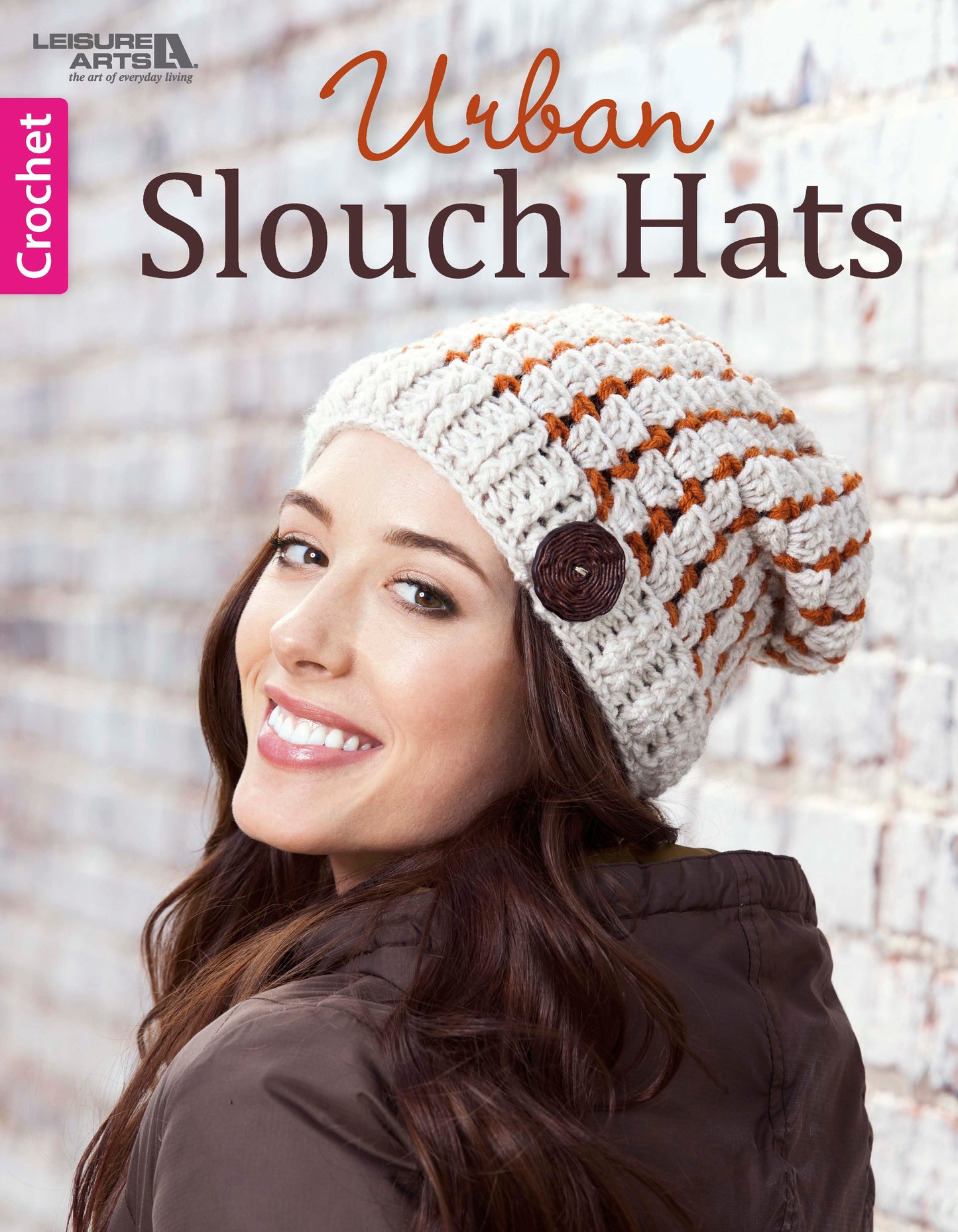 A smiling woman wears a white and orange striped Crochet Urban Slouch Hat by Leisure Arts, featuring a brown button. She stands before a white brick wall. Text reads: Crochet Urban Slouch Hats – Leisure Arts.