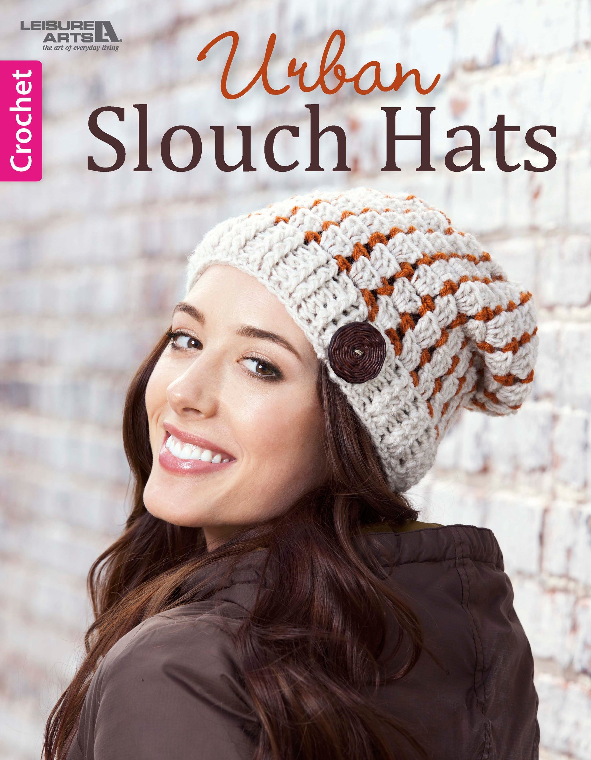 A smiling woman wears a white and orange striped Crochet Urban Slouch Hat by Leisure Arts, featuring a brown button. She stands before a white brick wall. Text reads: Crochet Urban Slouch Hats – Leisure Arts.