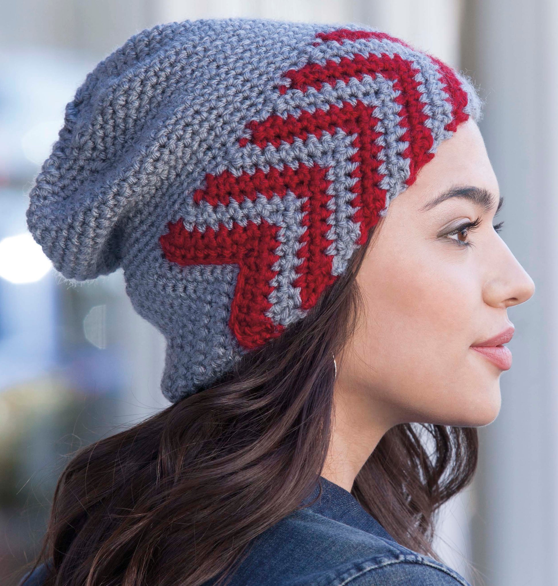 A woman in profile wears a Crochet Urban Slouch Hat by Leisure Arts, featuring light gray medium weight yarn with bold red zigzag patterns, set against a blurred outdoor background—ideal for fans of unique crochet styles.