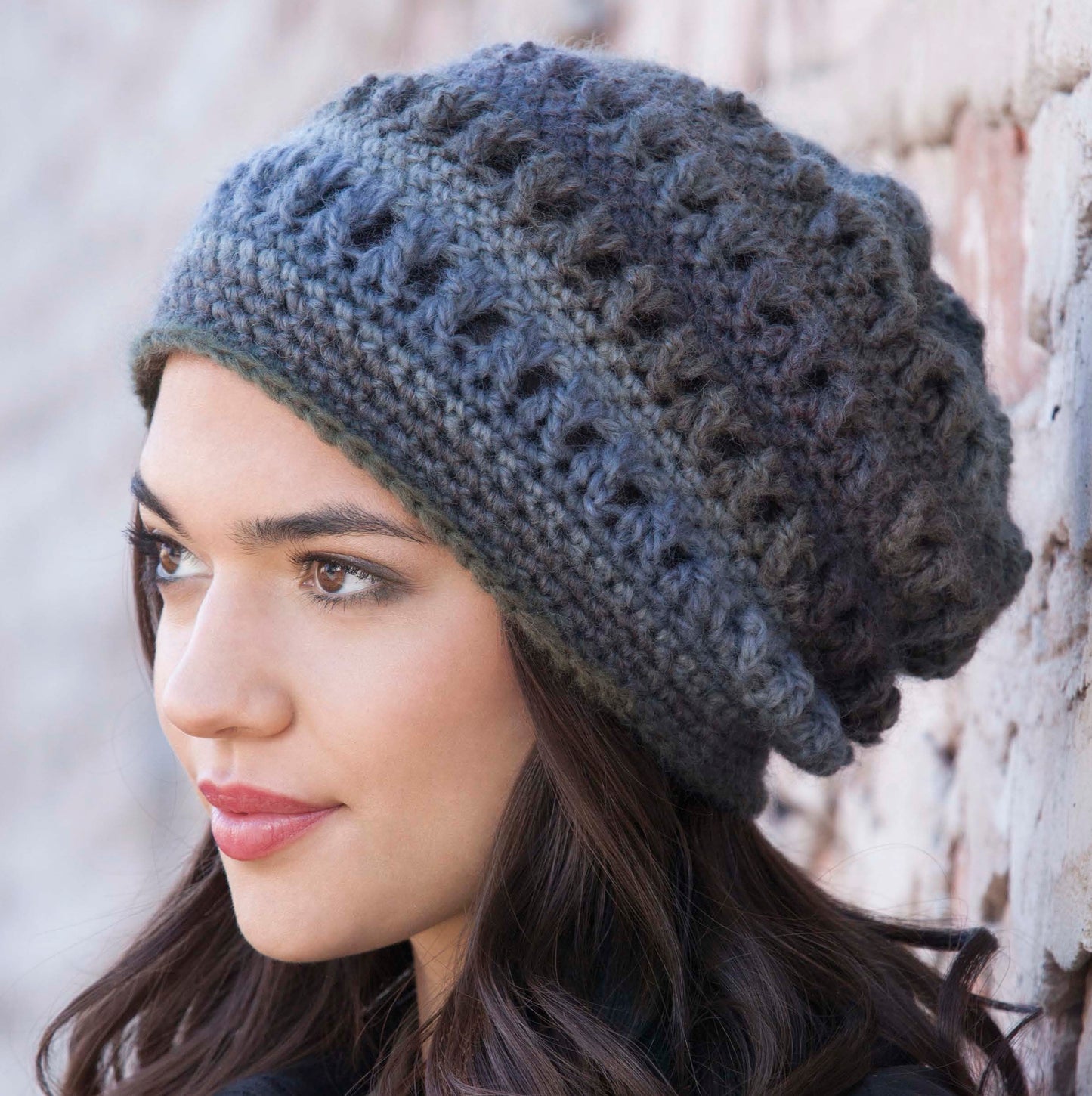 A woman with long dark hair wears a Leisure Arts Crochet Urban Slouch Hat in textured dark gray, made from medium weight yarn, as she looks to the side against a light-colored brick wall.