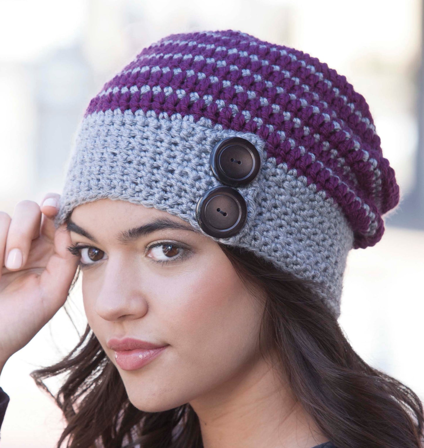 A woman with long dark hair wears the Crochet Urban Slouch Hat by Leisure Arts in gray and purple, featuring two large black buttons on the side. She gently touches the edge of the medium weight yarn beanie, gazing slightly past the camera.