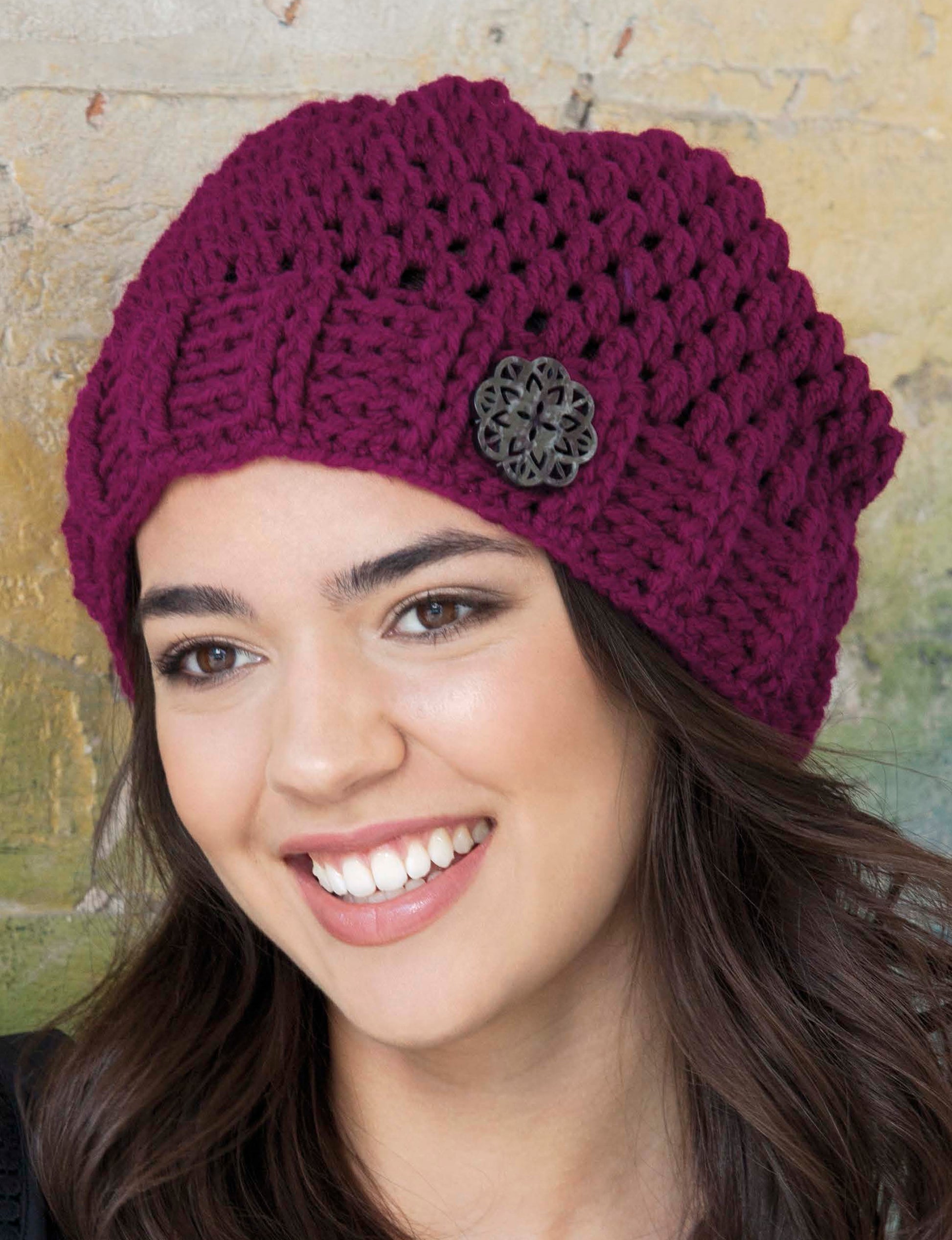 A person with long brown hair wears a Leisure Arts Crochet Urban Slouch Hat, a dark purple, medium-weight knit accessorized with an ornate, round button, standing before a textured yellow backdrop.