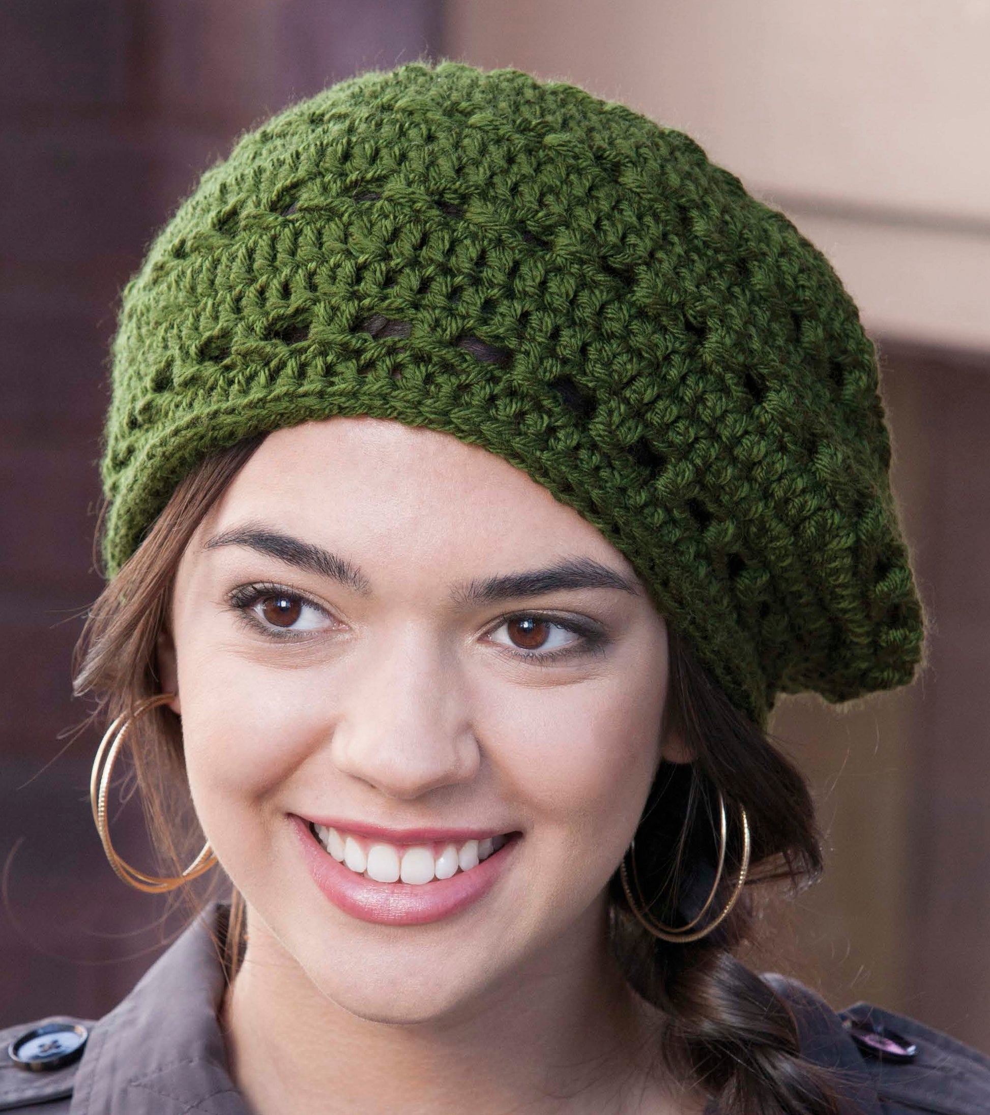 A smiling person models a green “Crochet Urban Slouch Hat” by Leisure Arts, styled loosely over brown hair and paired with large hoop earrings—great inspiration for beginner crocheters seeking fresh project ideas.