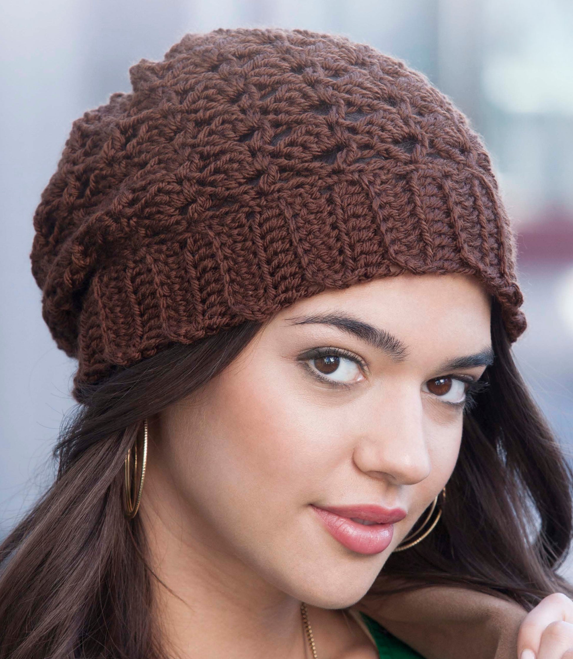 A woman with long dark hair wears a brown Crochet Urban Slouch Hat by Leisure Arts and gold hoop earrings, looking at the camera with a slight smile against a softly blurred background.