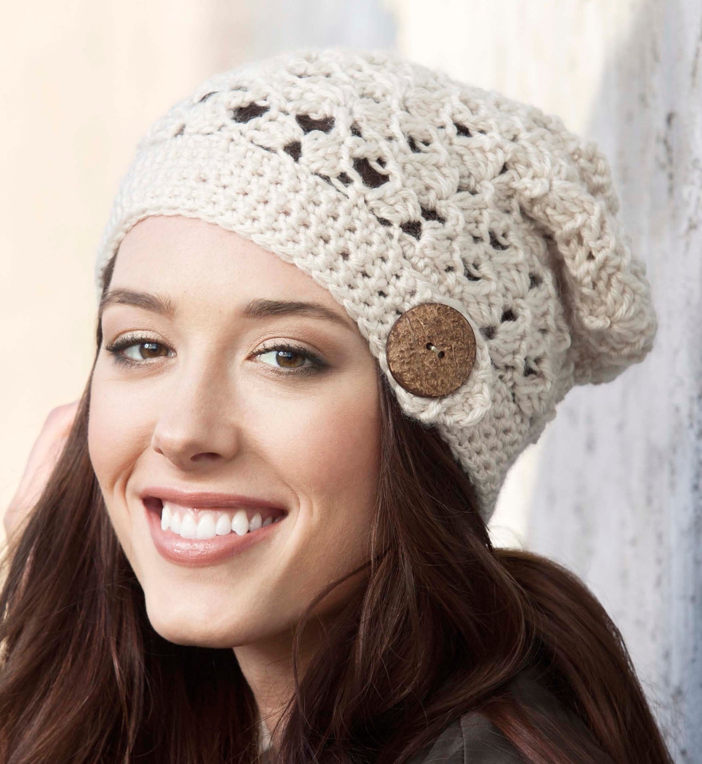 A smiling woman with long brown hair wears the Leisure Arts Crochet Urban Slouch Hat, a cream-colored, crocheted beanie with a large brown side button. She looks slightly left against a softly blurred background.