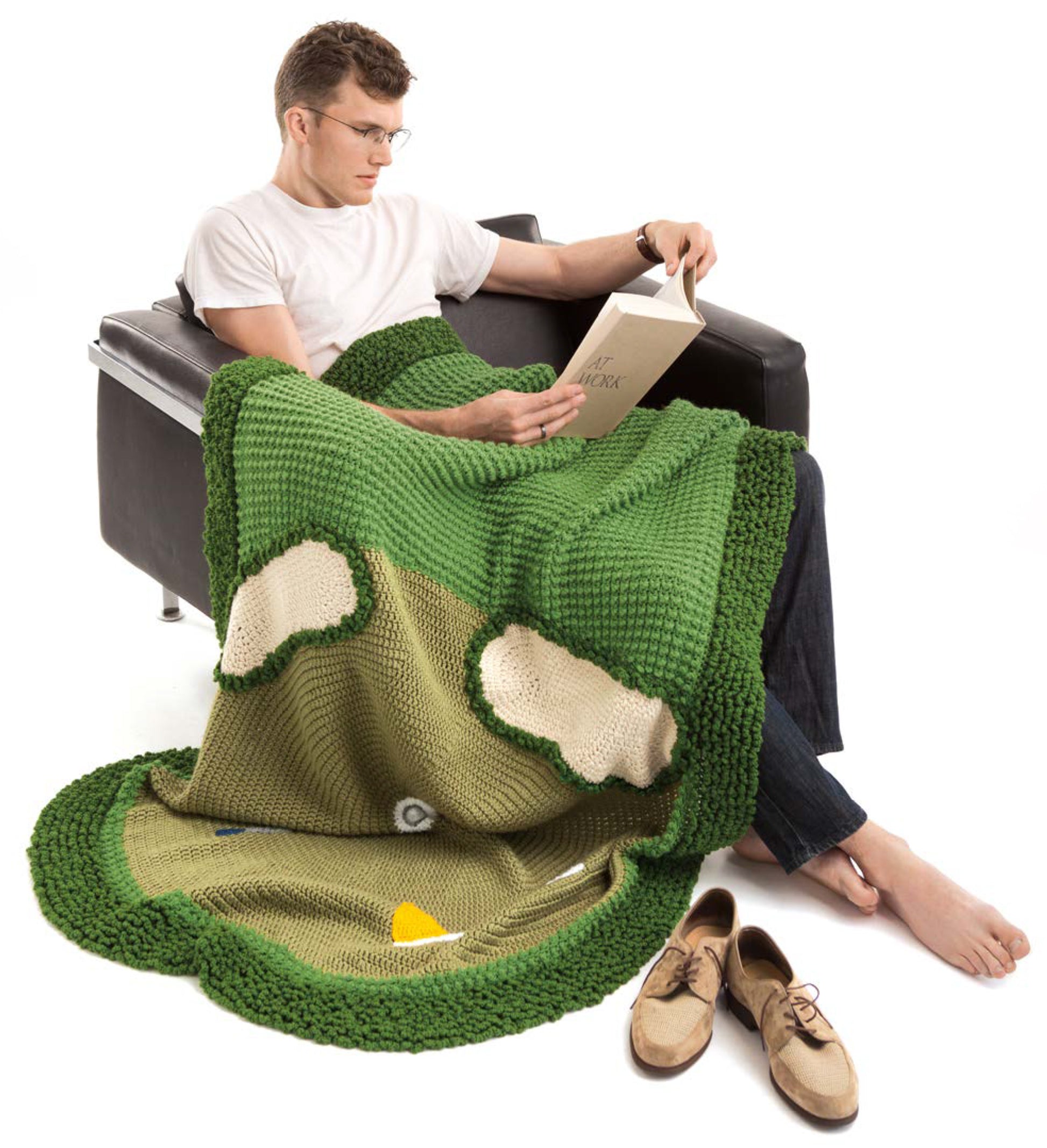 A man relaxes in a black armchair, reading with his shoes off and covered by a green golf course-themed afghan from Leisure Arts' "Sportacular Afghans to Crochet" Digital Download—ideal man cave decor for any sports fan.