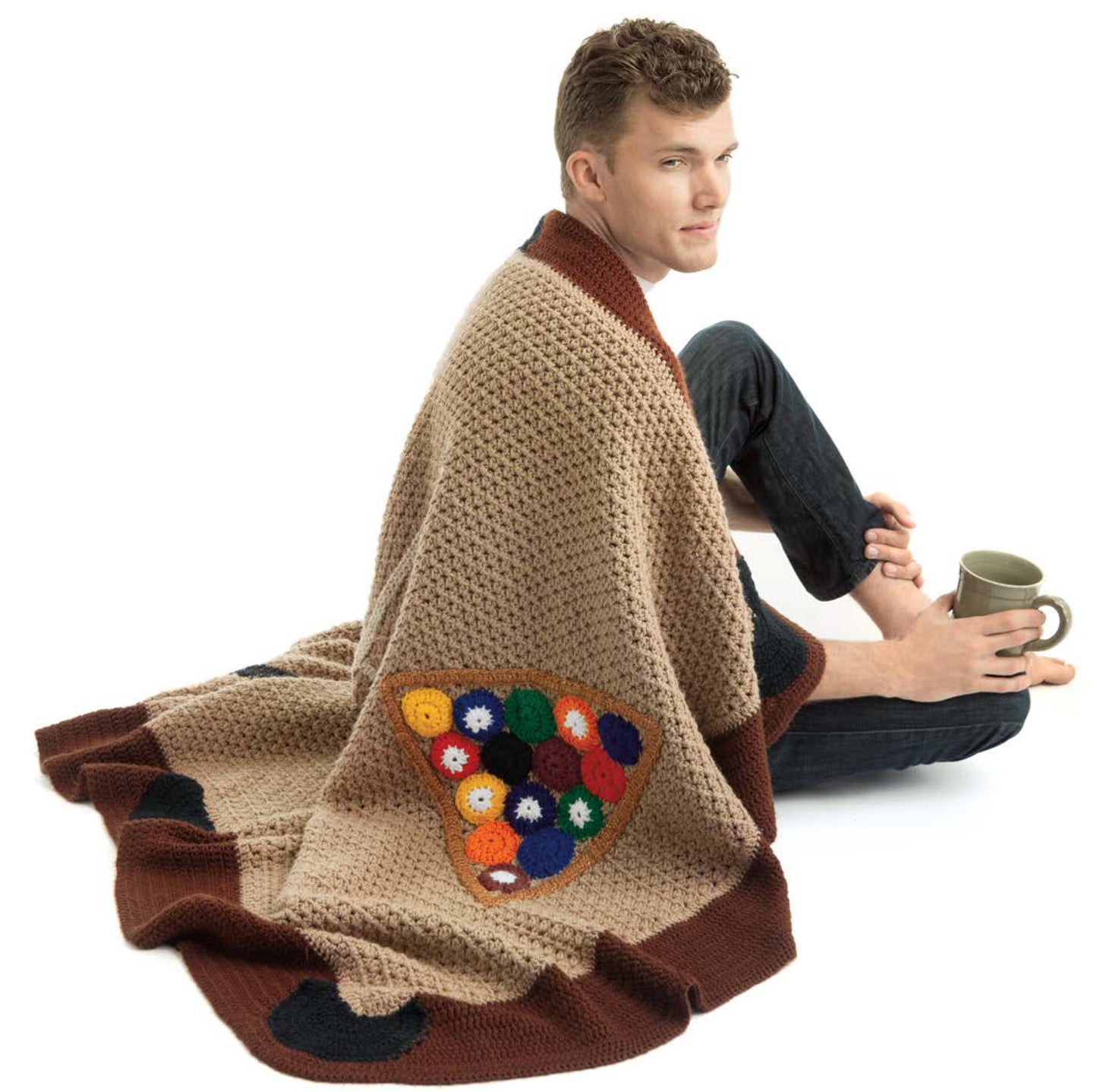 A man sits on the floor with a mug, wrapped in a beige crochet blanket featuring colorful accents—a perfect touch of unique style from the "Sportacular Afghans to Crochet Digital Download" by Leisure Arts for your man cave.